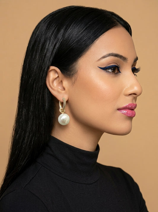 A woman in profile wearing a gold hoop earring with a dangling white pearl, set against a solid warm beige background.