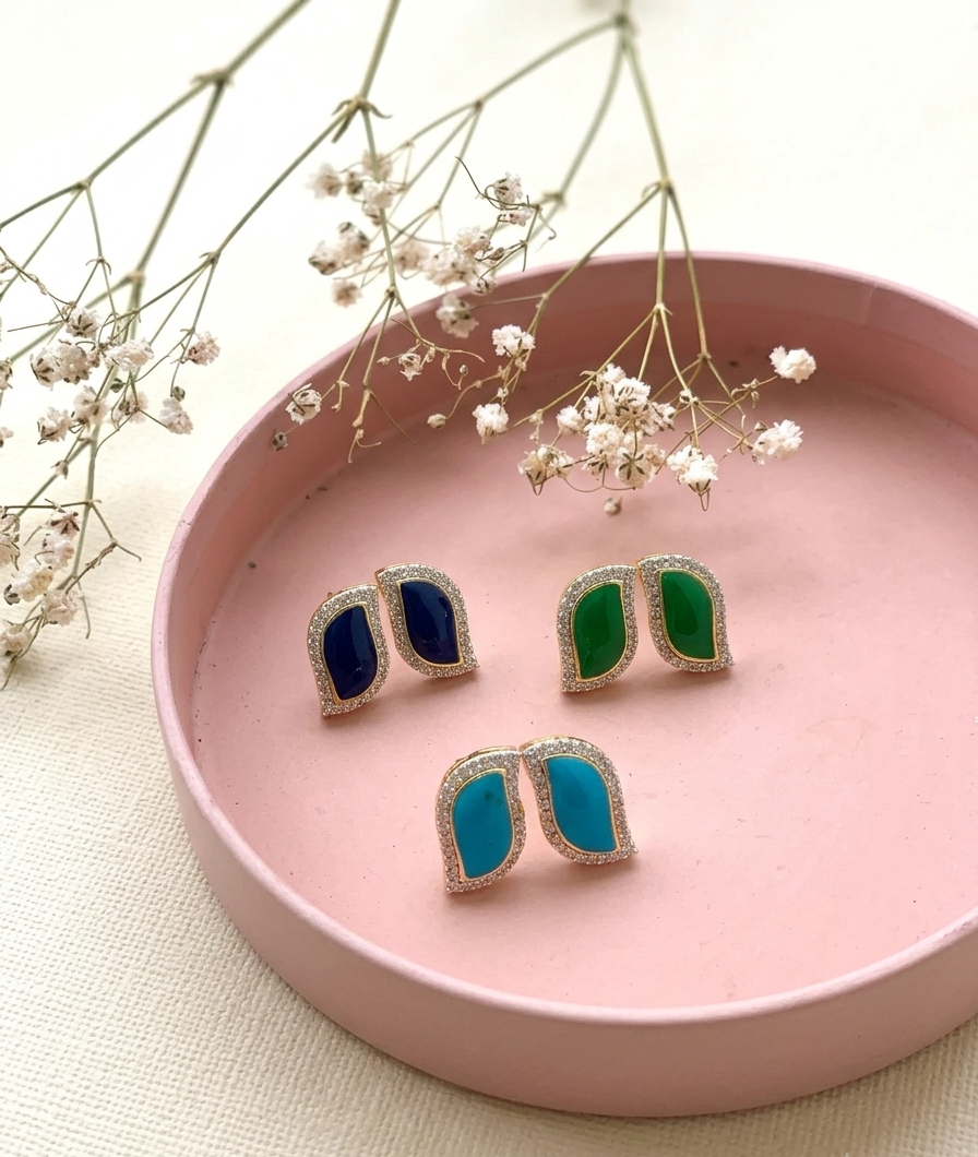 Three pairs of gold-toned stud earrings with curved, shield-like shapes and colored inlay stones in navy blue, emerald green, and turquoise, arranged on a round pink tray with dried baby's breath flowers.