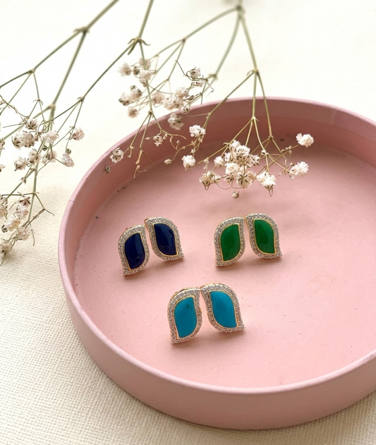 Three pairs of gold-toned stud earrings with curved, shield-like shapes and colored inlay stones in navy blue, emerald green, and turquoise, arranged on a round pink tray with dried baby's breath flowers.