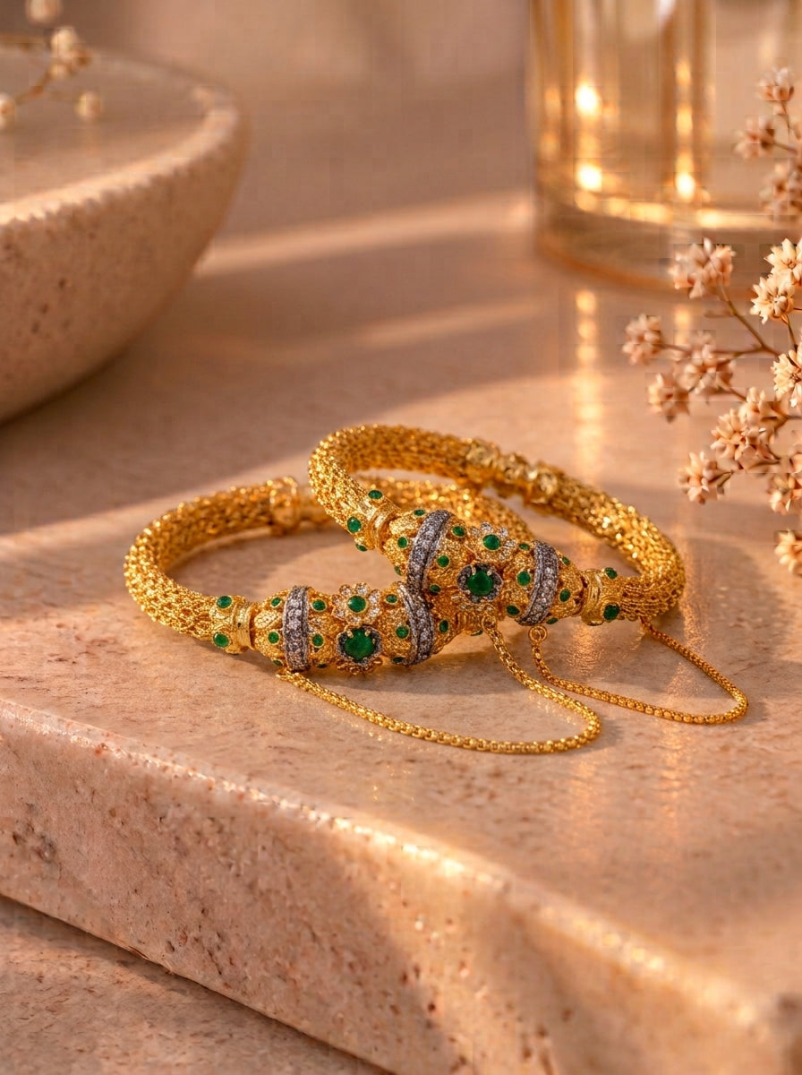 Two gold-plated bangles with green and silver stone embellishments, displayed on a beige stone surface with warm, soft-focus background props.