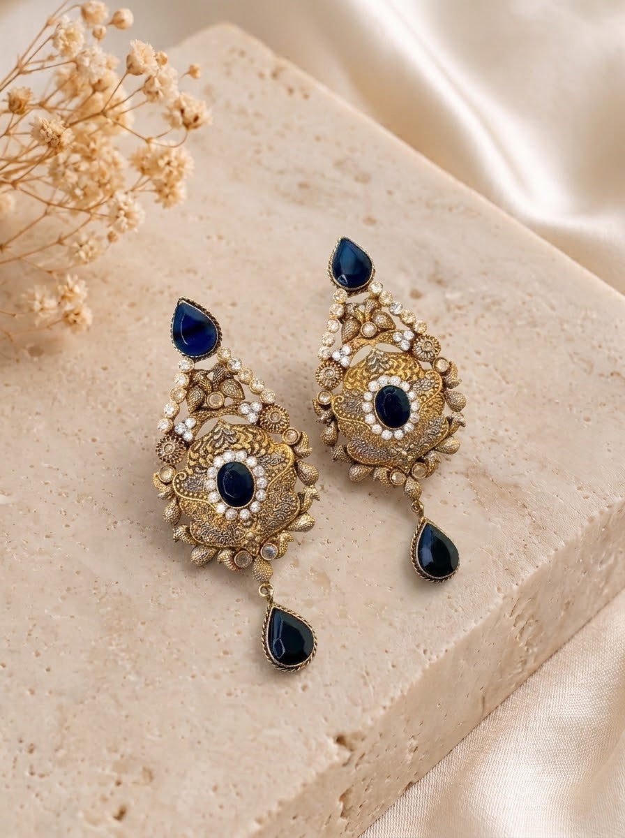 A pair of ornate gold-toned statement earrings with dark blue teardrop gemstones and white pearl-like accents, displayed on a beige stone block with dried flowers and satin fabric.