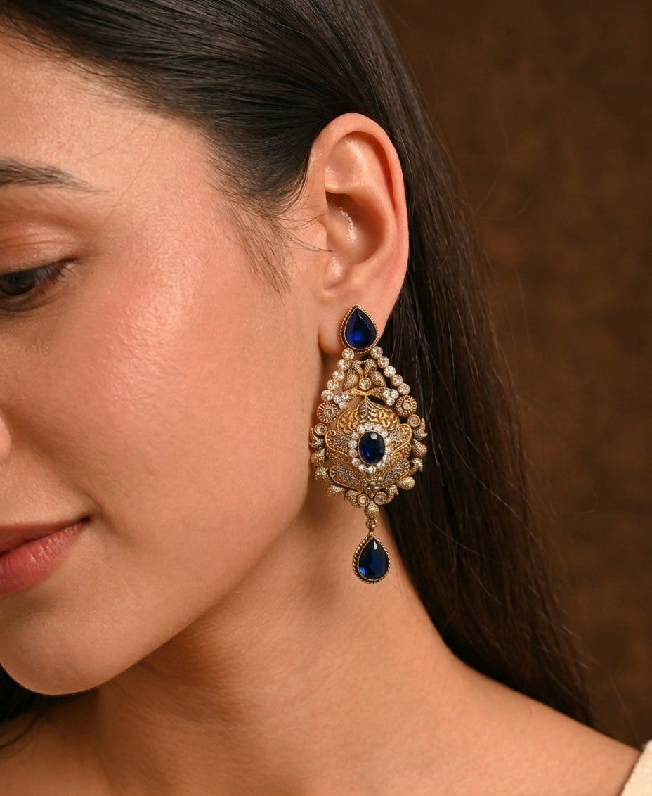 A close-up of a woman wearing an ornate gold-toned dangle earring with deep blue stones and small clear stones, shown against a warm brown background.