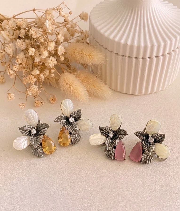 Four decorative flower-shaped earrings arranged on a light beige surface, with dried flowers and a ribbed white container in the background. The earrings feature silver-toned leaf settings, white iridescent petals, clear rhinestone accents, and teardrop gemstones in yellow and pink.