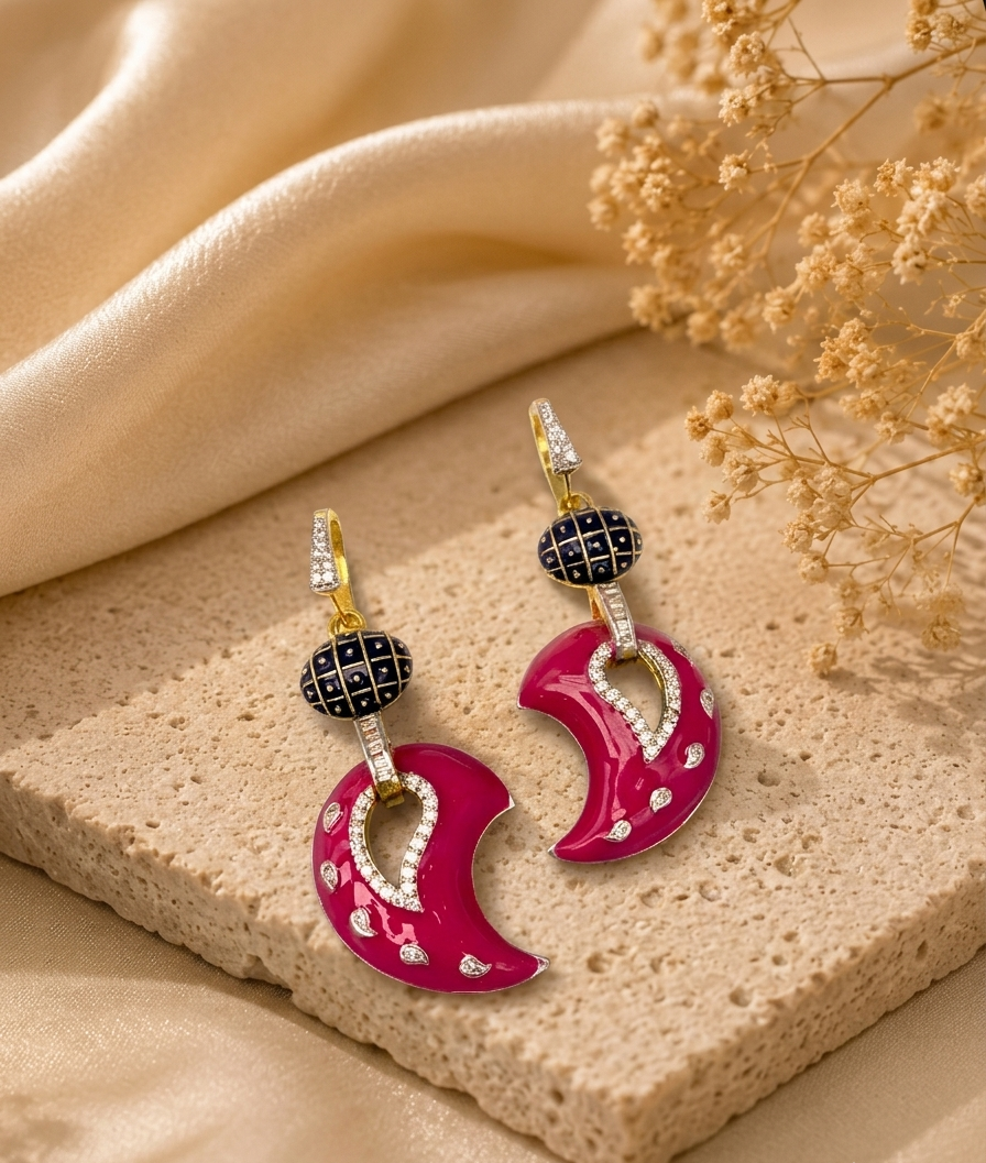 A pair of gold-tone drop earrings with bright pink crescent moon charms, black patterned beads, and small clear stones, styled on a beige stone slab with cream fabric and dried flowers.