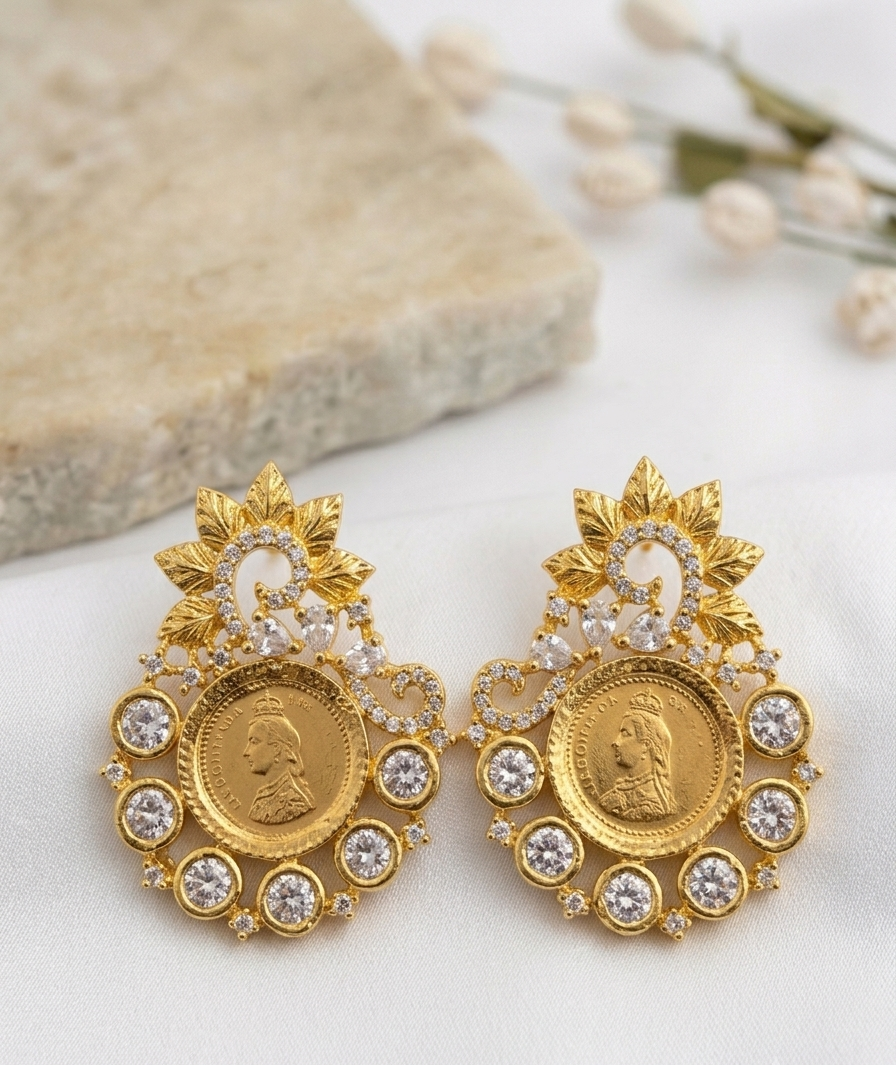 A pair of ornate gold-toned statement earrings with circular medallion motifs, floral leaf tops, and clear rhinestone accents, displayed on a light surface with a stone and dried flowers in the background.