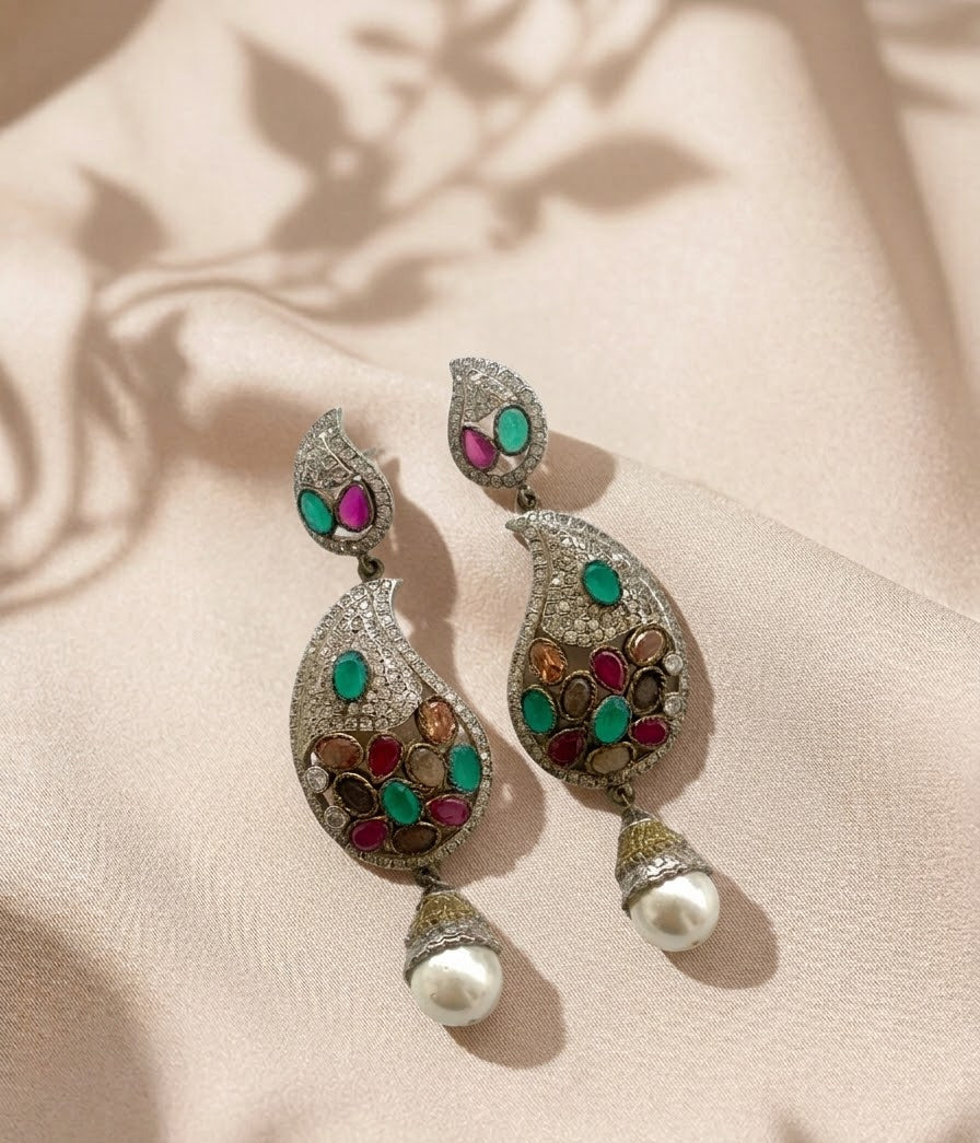 A pair of ornate silver-toned Indian-style dangle earrings with colorful inlay stones and white pearl drops, displayed on a beige satin fabric with leaf shadows.
