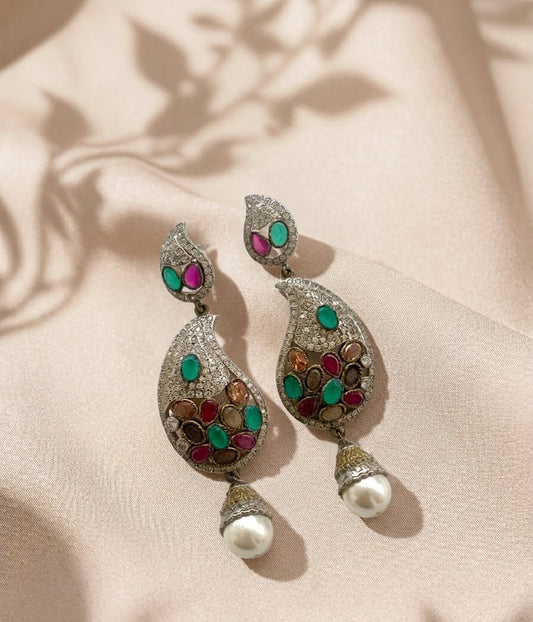 A pair of ornate silver-toned Indian-style dangle earrings with colorful inlay stones and white pearl drops, displayed on a beige satin fabric with leaf shadows.