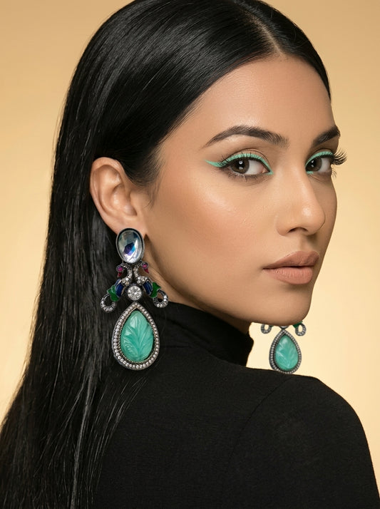 A close-up portrait of a woman with long black hair wearing large statement earrings. The earrings feature prominent turquoise-colored teardrop stones with silver-toned metal settings and smaller multicolored accents.
