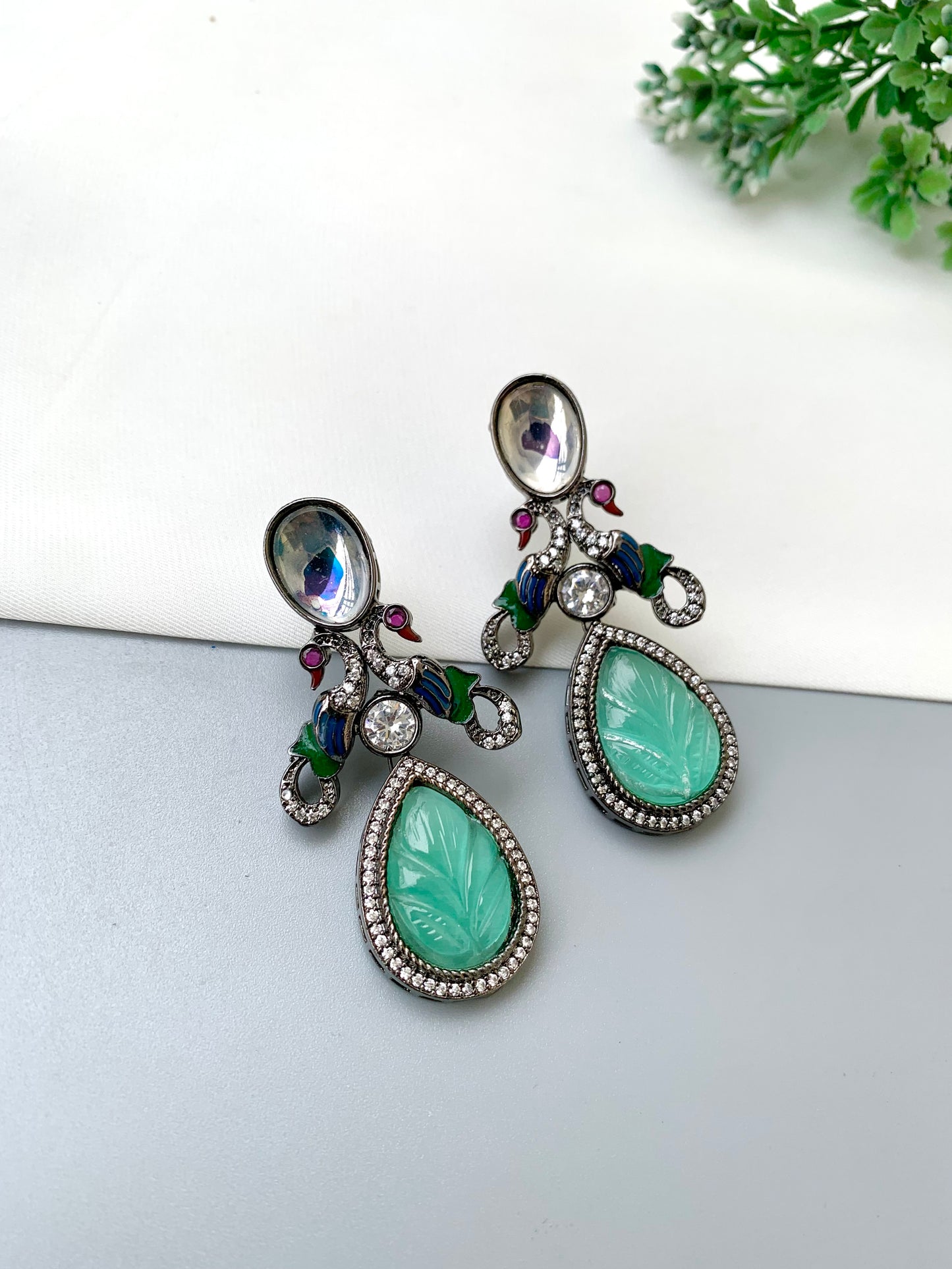 A pair of ornate silver-toned dangle earrings with large teardrop-shaped light green carved stone centers, surrounded by clear rhinestones and small green and purple enamel accents, displayed on a light gray surface.