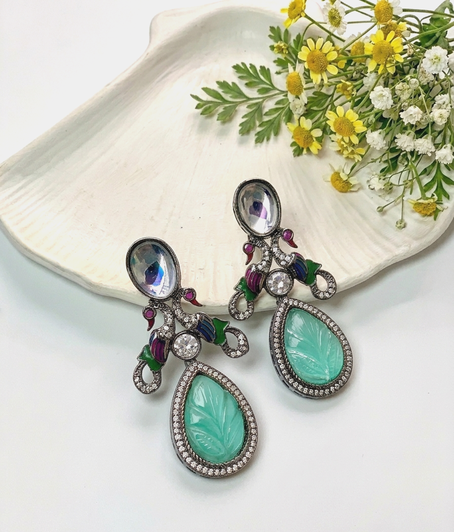 A pair of ornate drop earrings with large mint green teardrop-shaped stones, silver-toned settings, clear rhinestone accents, and small purple and green floral details, displayed on a white shell-shaped tray with yellow and white flowers.