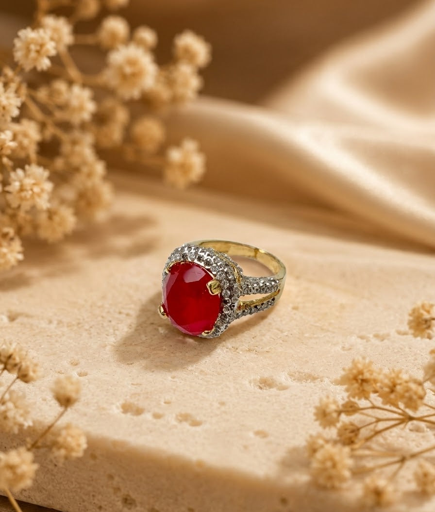 A gold ring with a large oval red gemstone and small clear stones along the band, styled on a beige stone surface with dried flowers and soft satin fabric in the background.