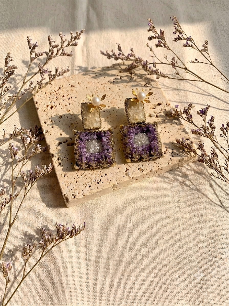 A pair of gold-toned statement earrings with square purple crystal clusters and small pearl accents, displayed on a speckled stone slab with dried flowers on a beige fabric background.