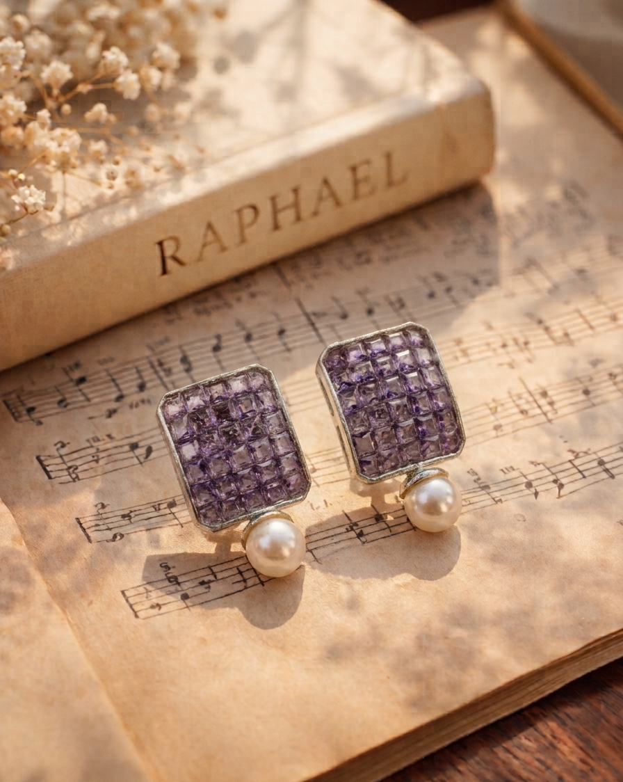 A pair of rectangular stud earrings with purple crystal grids and white pearl drops, displayed on an open vintage book over sheet music with a book titled RAPHAEL in the background.