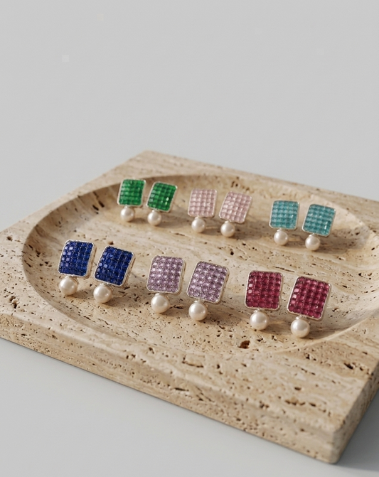 Six pairs of stud earrings displayed on a beige travertine tray against a light gray background. Each pair features a small square grid of rhinestones in a different color—green, pink, blue, lavender, teal, and magenta—terminated by a single white pearl stud.