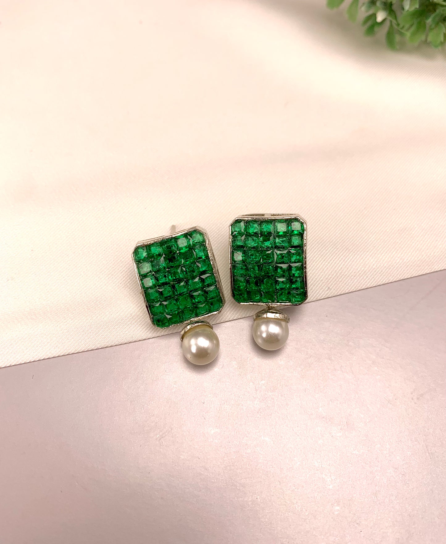 A pair of square stud earrings featuring a textured green crystal top and a dangling white pearl, displayed on a light pink textured surface.