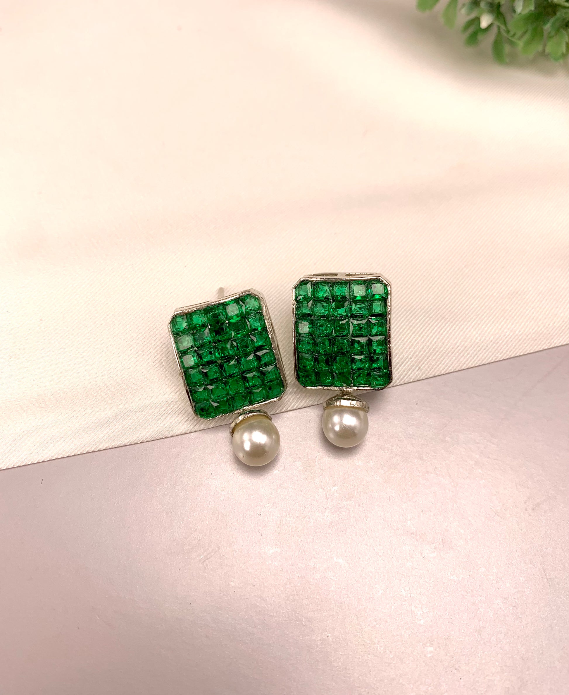 A pair of square stud earrings featuring a textured green crystal top and a dangling white pearl, displayed on a light pink textured surface.