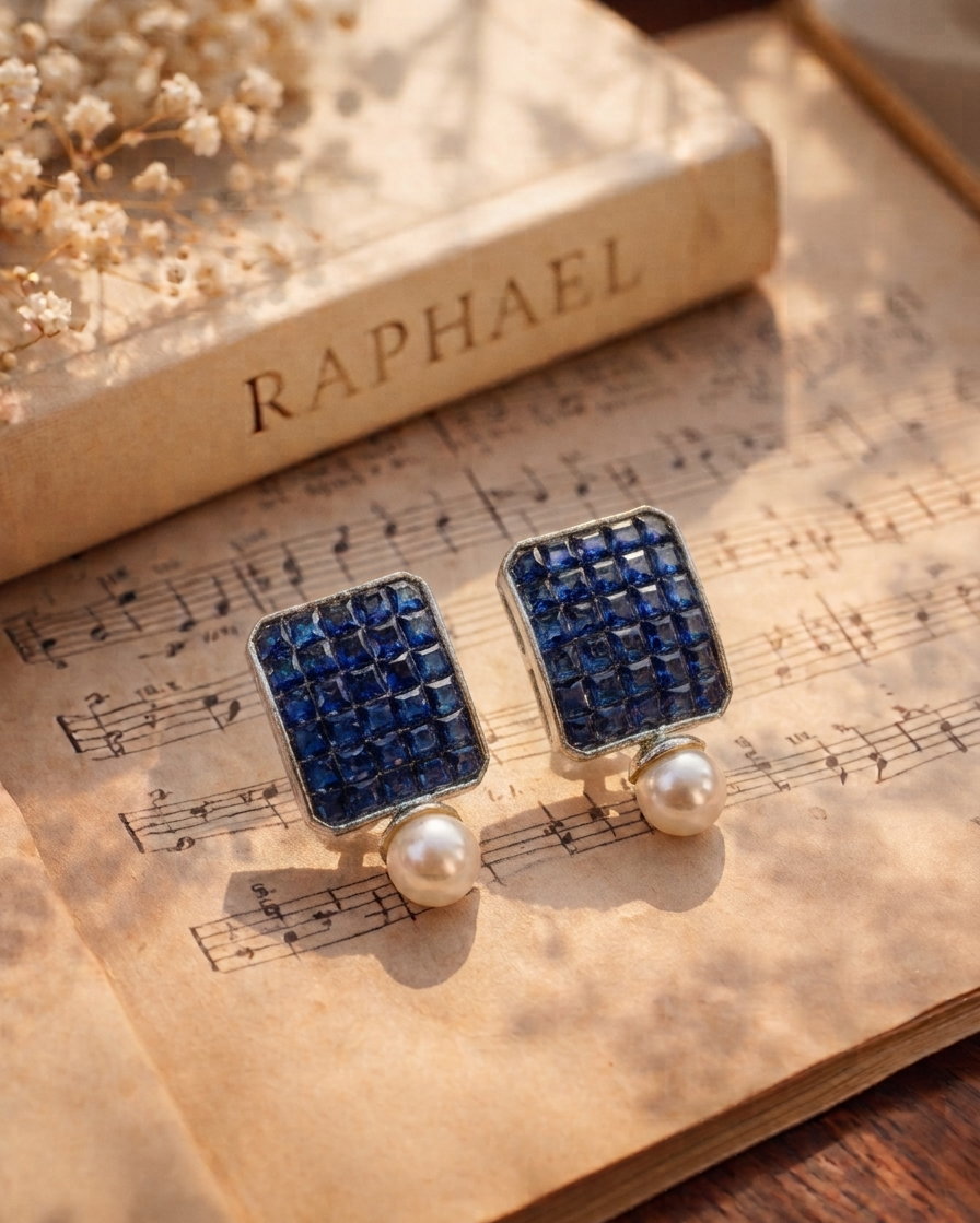 A pair of rectangular stud earrings with deep blue rhinestone grids and white pearl drops, displayed on an open vintage book with sheet music and dried flowers. The book spine in the background is labeled 'RAPHAEL'.