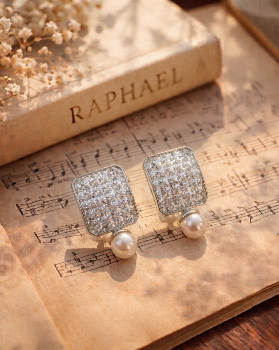 A pair of rectangular silver-toned earrings paved with clear stones and topped with white pearl drops, displayed on an open book of sheet music next to a vintage book labeled RAPHAEL.