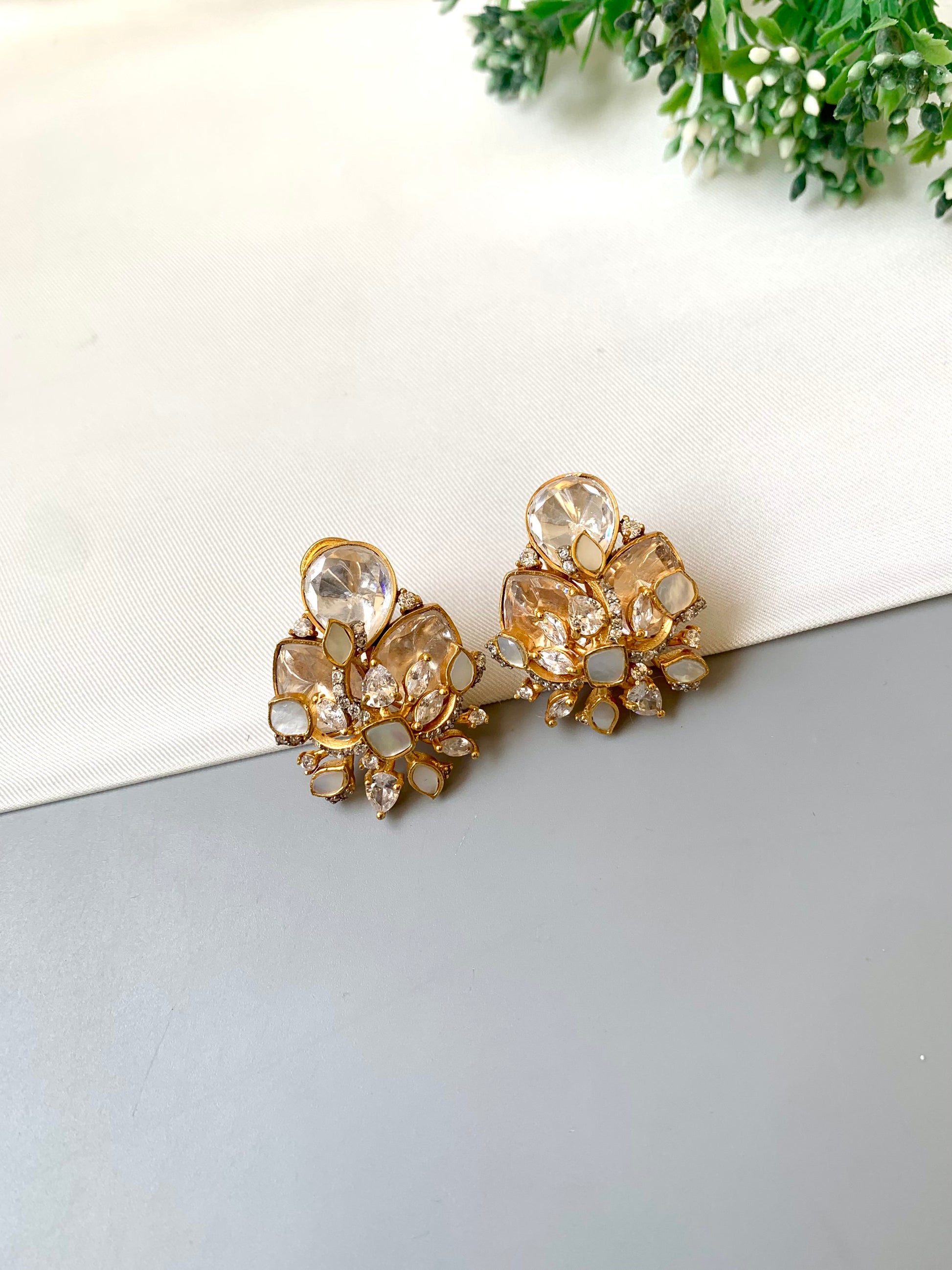 Gold floral earrings with clear stones on a light gray surface with greenery in the background.