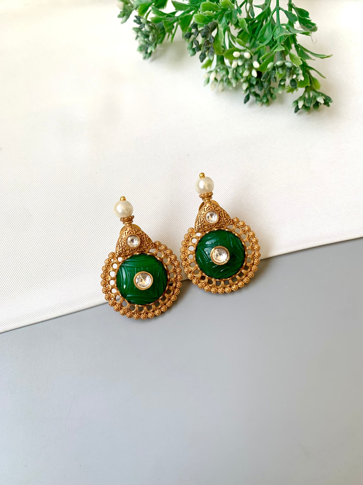 Gold and green earrings on a white surface with green leaves in the background