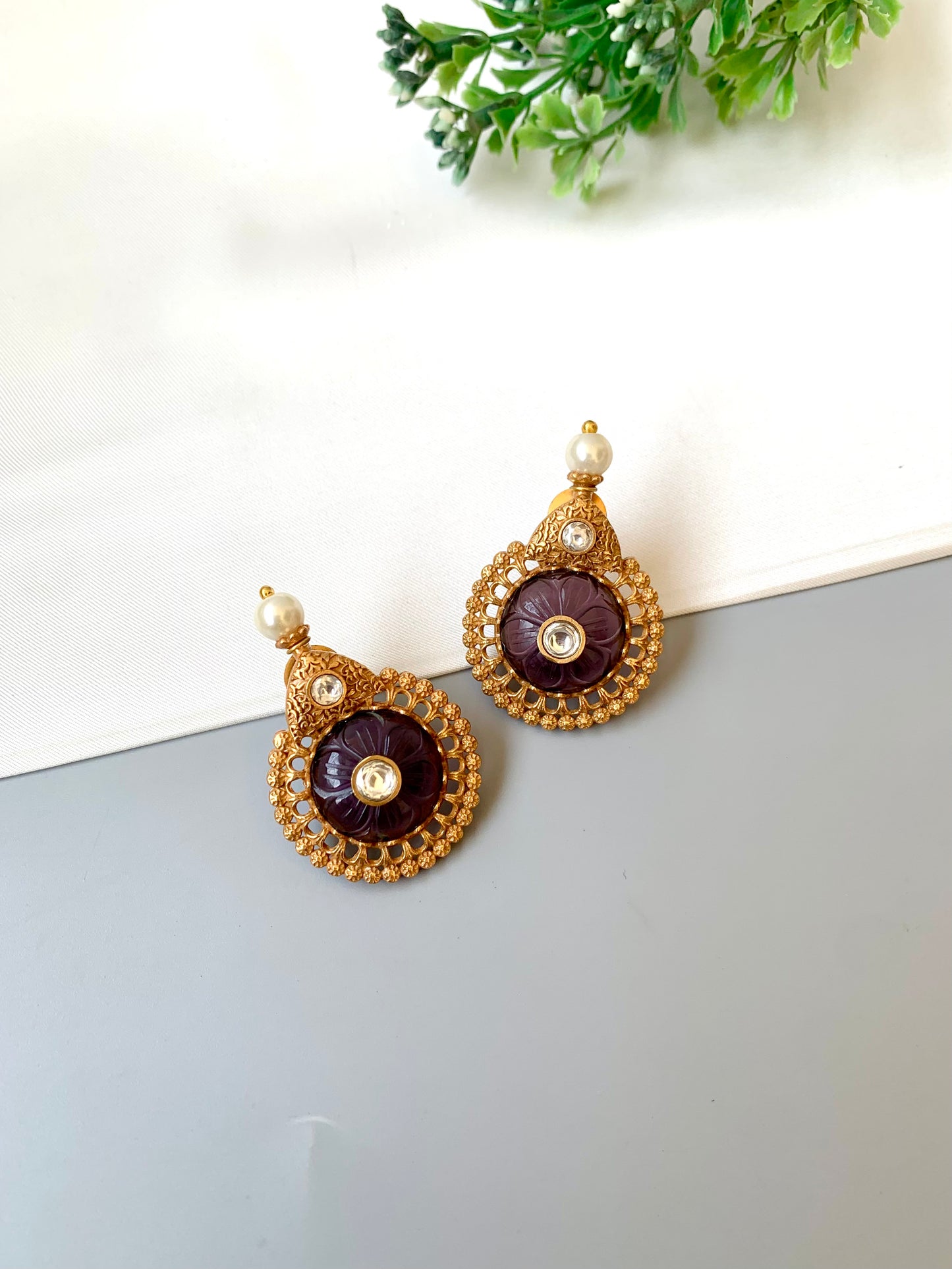 Pair of gold and purple earrings on a white surface with green leaves in the background