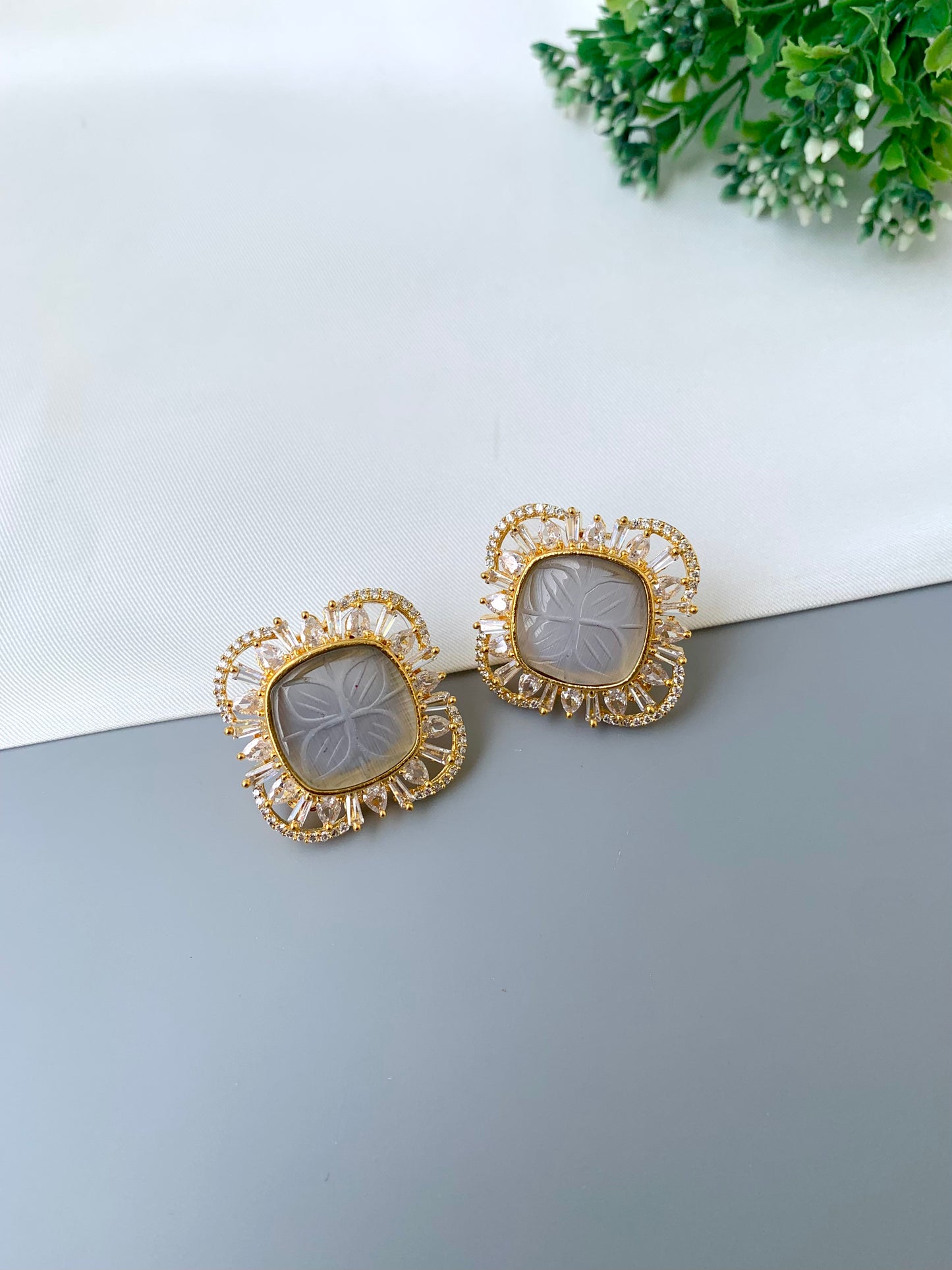 Gold-framed earrings on a gray surface with a white background and greenery.