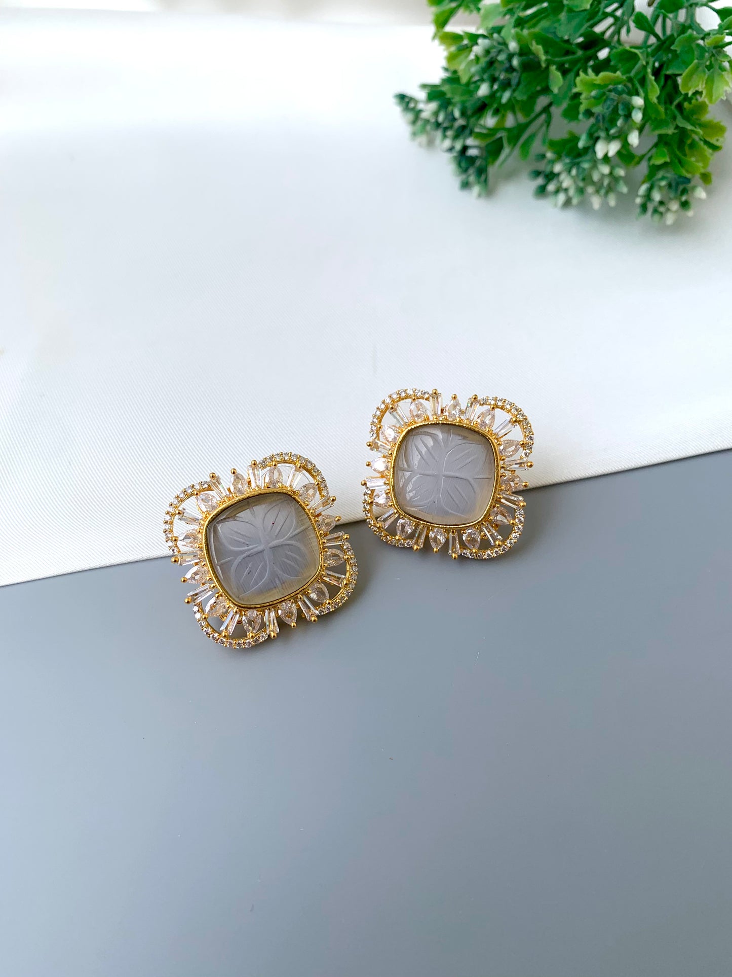 Gold earrings with intricate designs on a light surface with greenery in the background