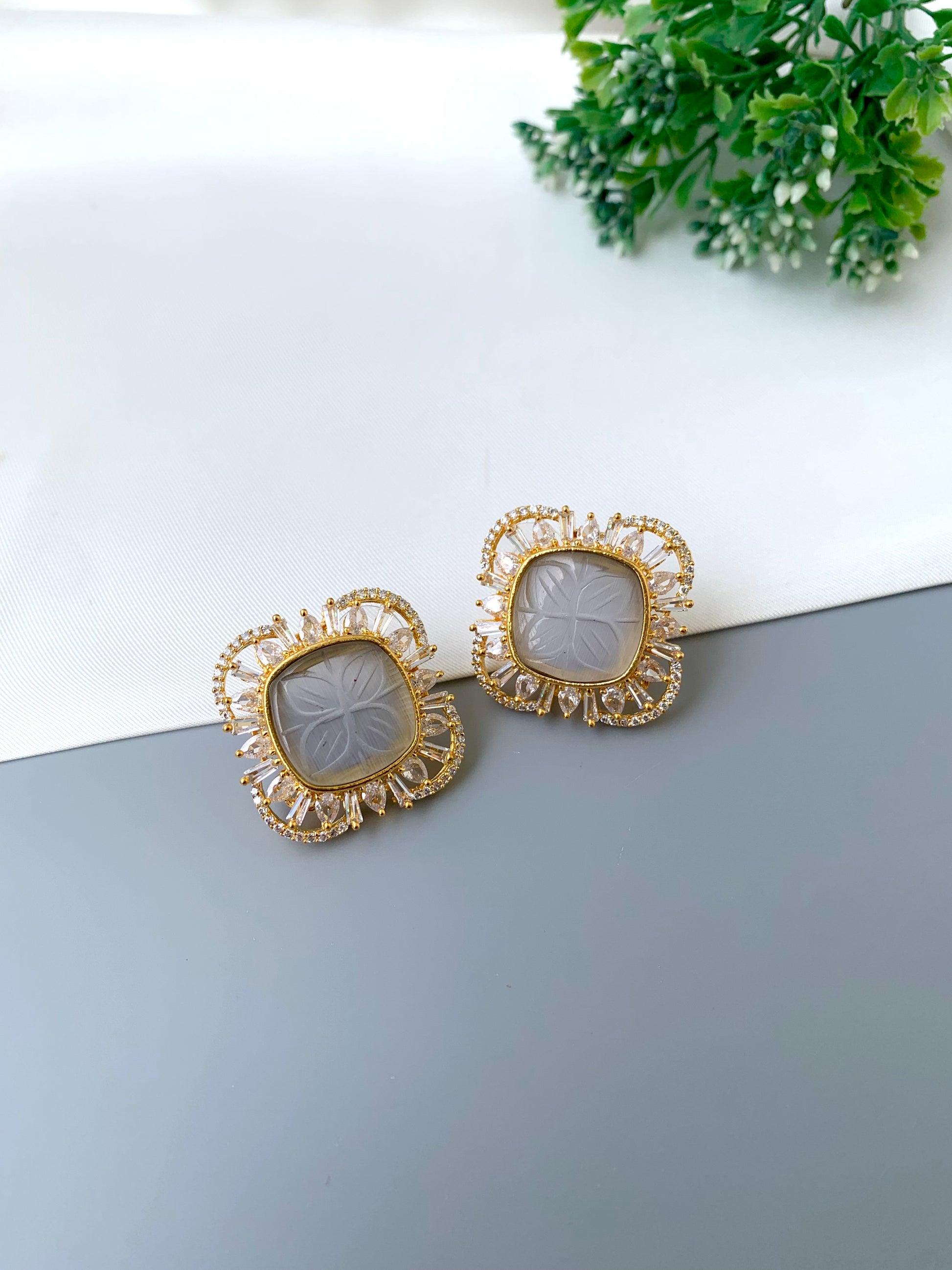 Gold earrings with intricate designs on a light surface with greenery in the background