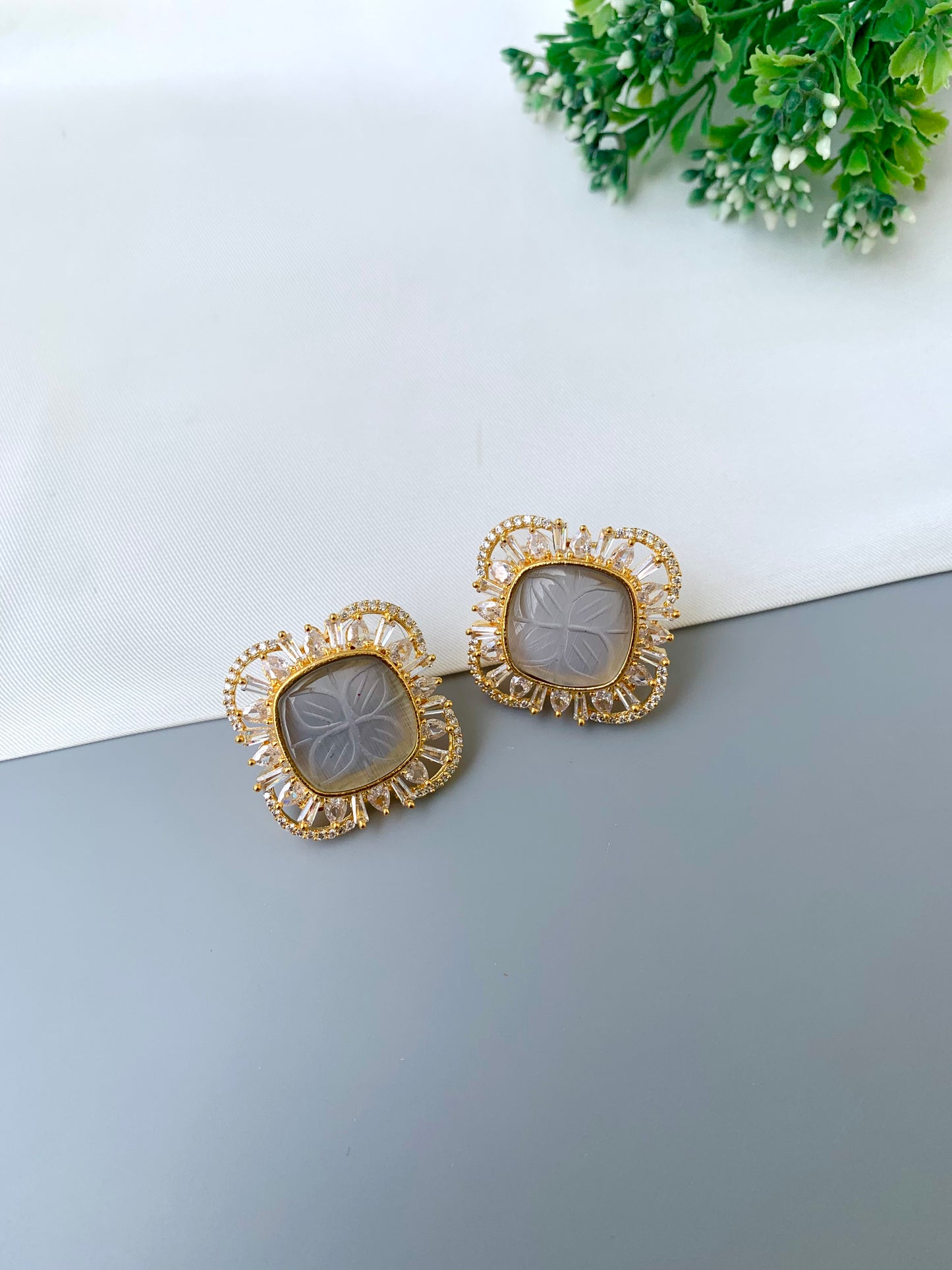 Gold earrings on a gray surface with a white background and greenery.