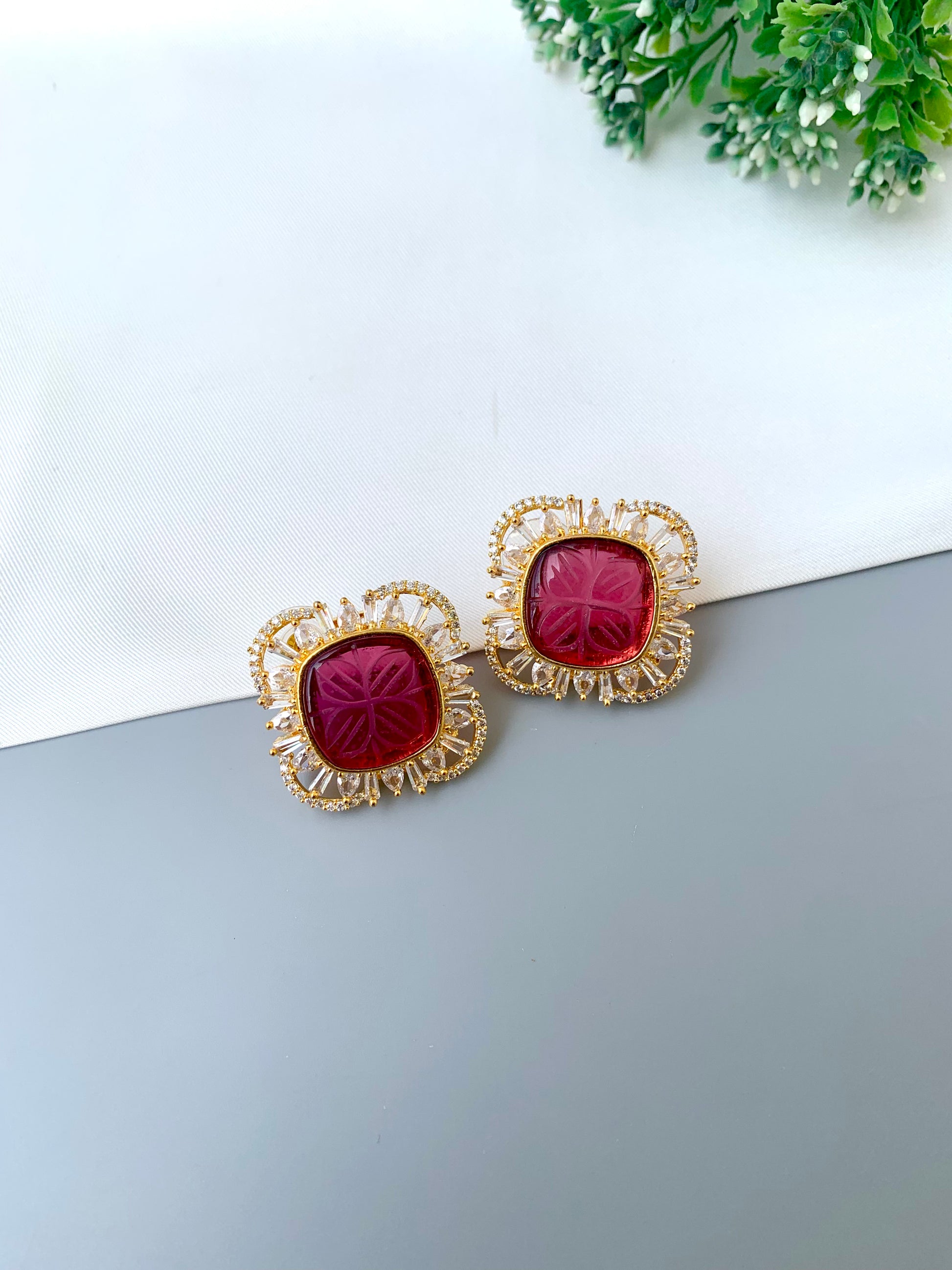 Gold earrings with red gemstones on a white surface with greenery in the background