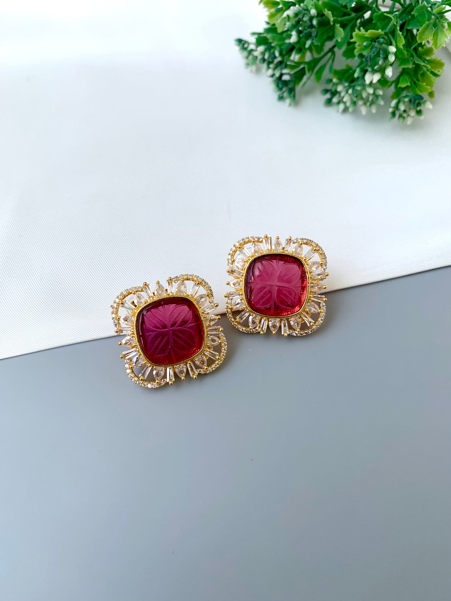 Gold earrings with red gemstones on a white surface with greenery in the background