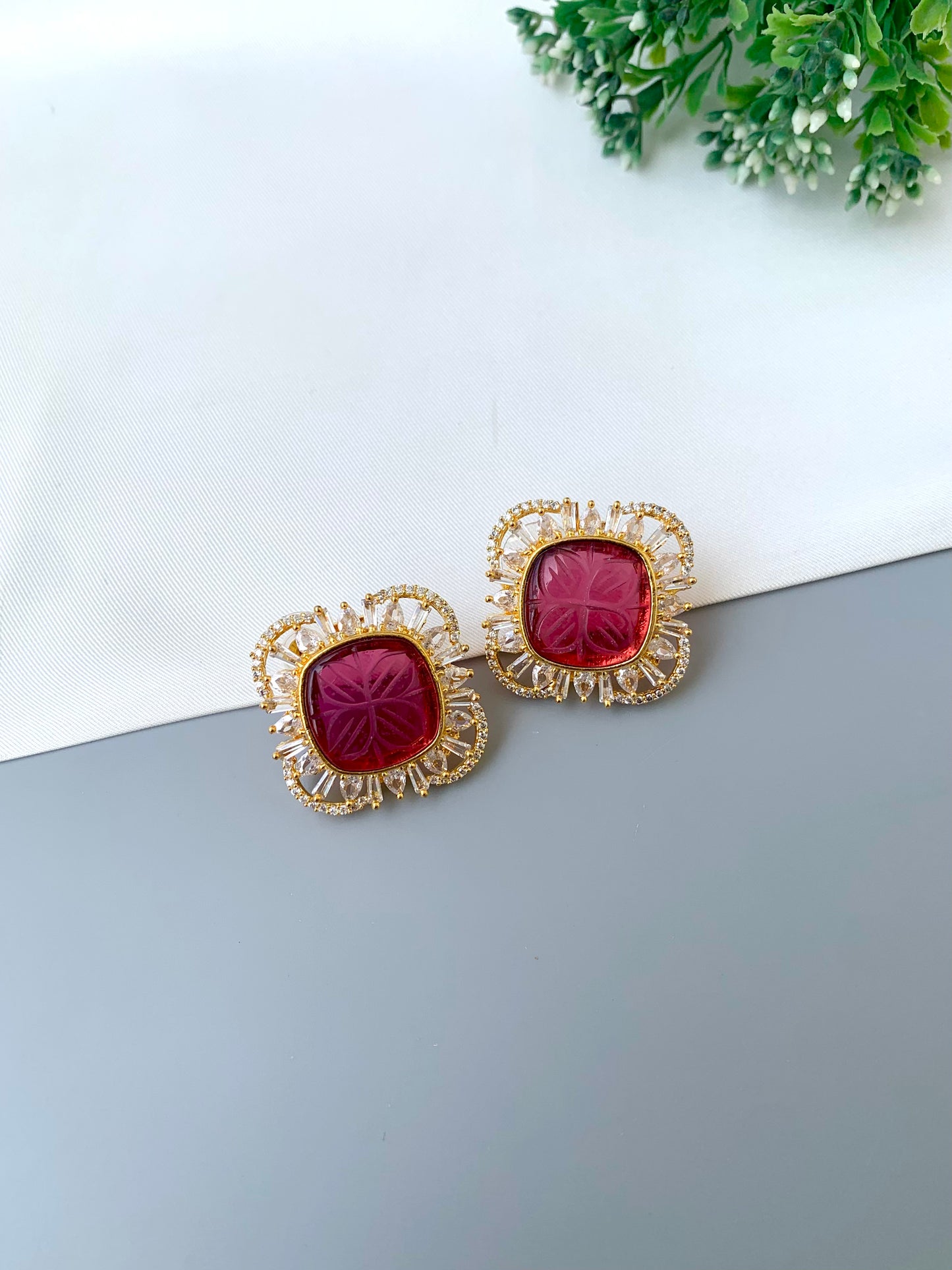 Gold earrings with red gemstones on a gray surface with greenery in the background