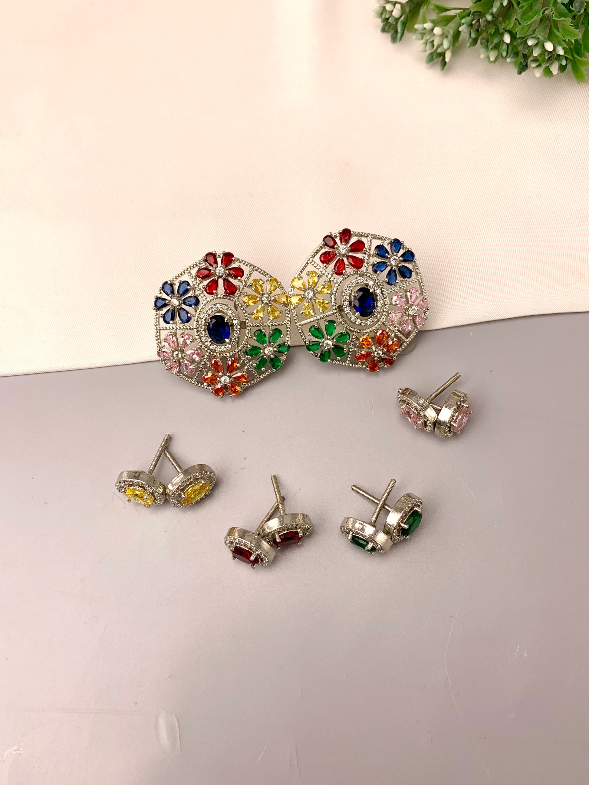 A set of silver-toned jewelry pieces displayed on a light gray surface against a cream background, including a pair of large floral stud earrings and several smaller matching stud earrings with colored gemstones.