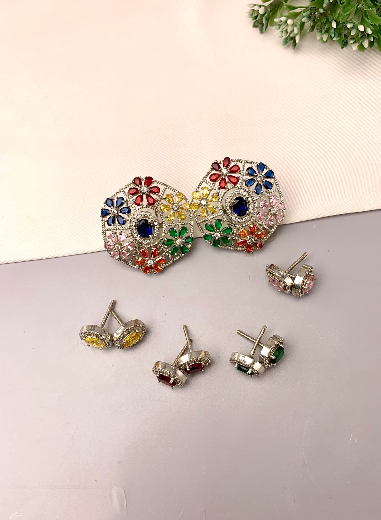 A pair of large silver-tone statement earrings with colorful floral enamel details, shown alongside four matching stud earrings on a light gray surface against a cream background.