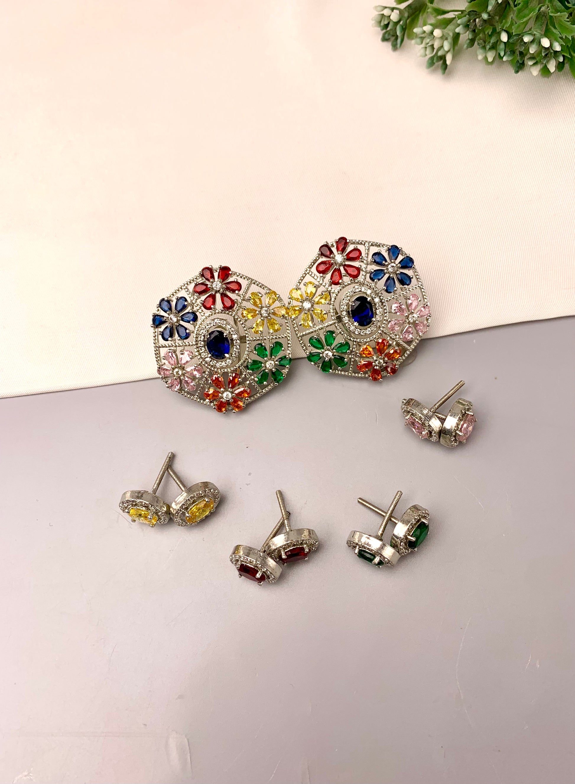 A pair of large silver-tone statement earrings with colorful floral enamel details, shown alongside four matching stud earrings on a light gray surface against a cream background.
