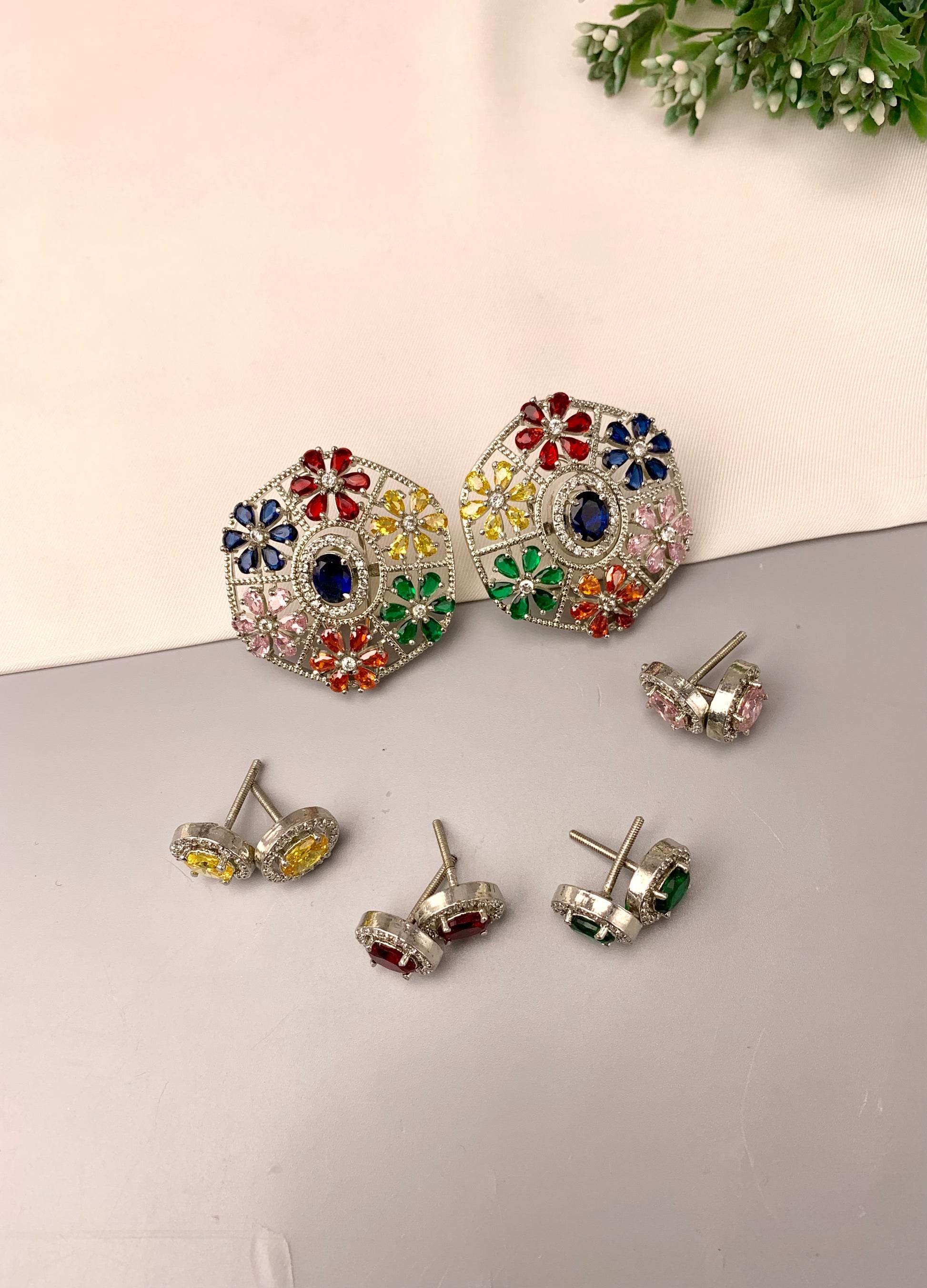 A set of silver-toned stud earrings displayed on a split beige and gray surface. The main pair features a large, colorful floral mandala design with blue, red, yellow, green, and pink stones in the center and around the border.