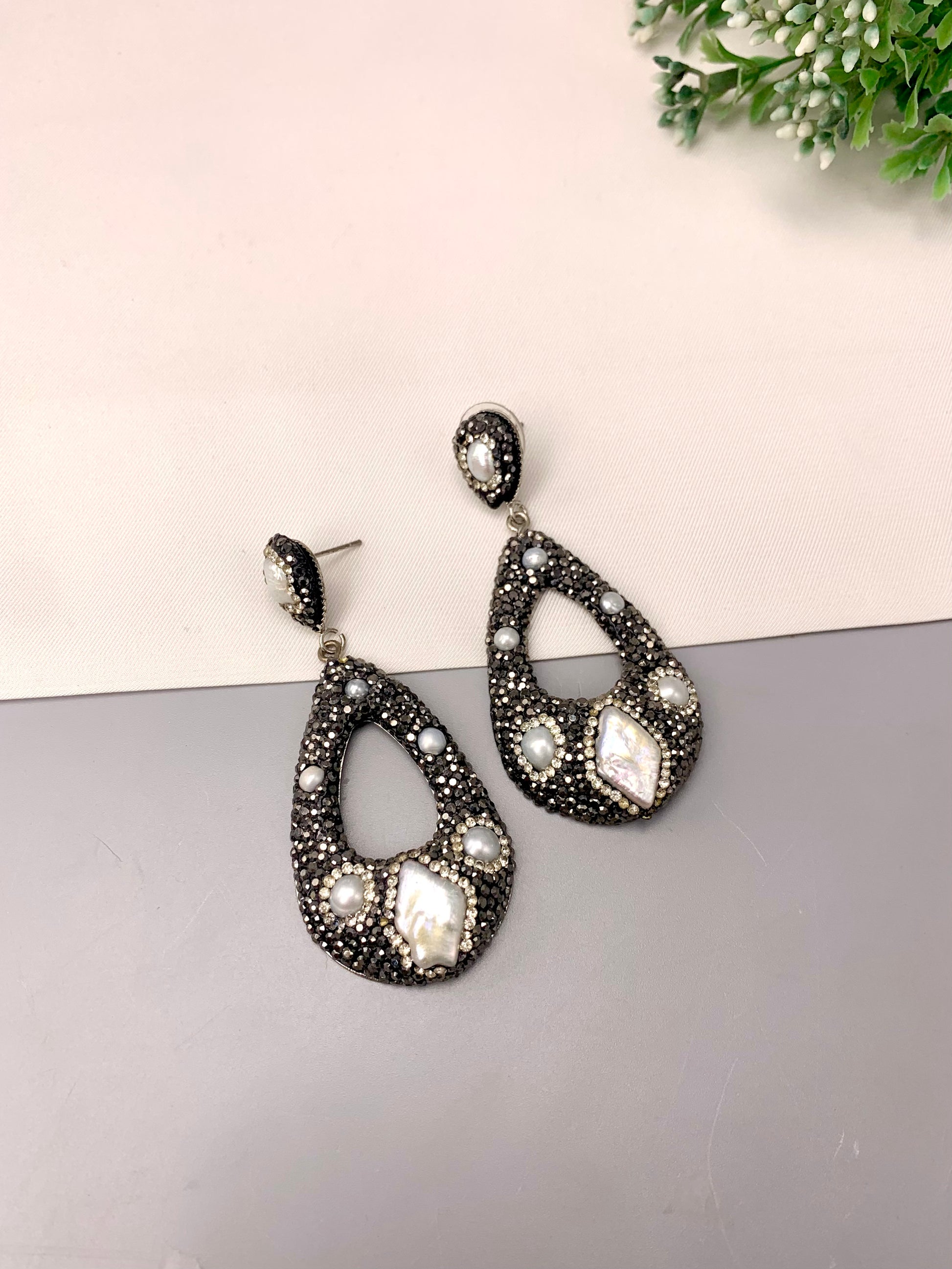 A pair of teardrop-shaped drop earrings with a black textured pavé setting, adorned with white faux pearls and a large white marquise-cut stone, displayed on a split white and gray surface.