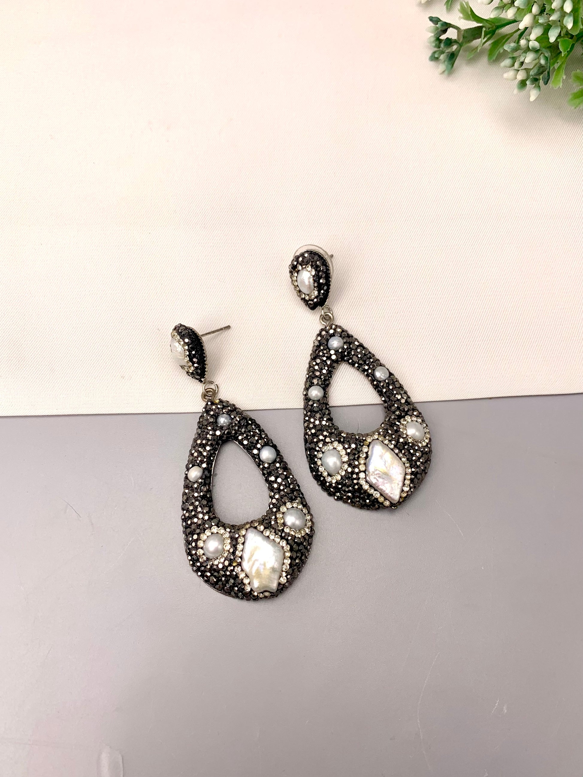 A pair of teardrop-shaped drop earrings with a black textured base, adorned with white faux pearls and clear rhinestones, displayed on a split white and gray surface.