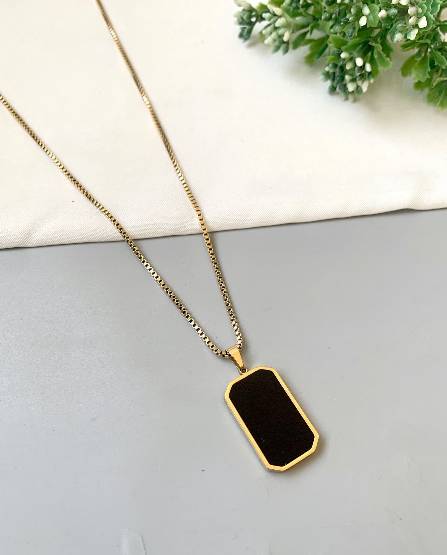 Gold necklace with a black pendant on a light gray surface with greenery in the background