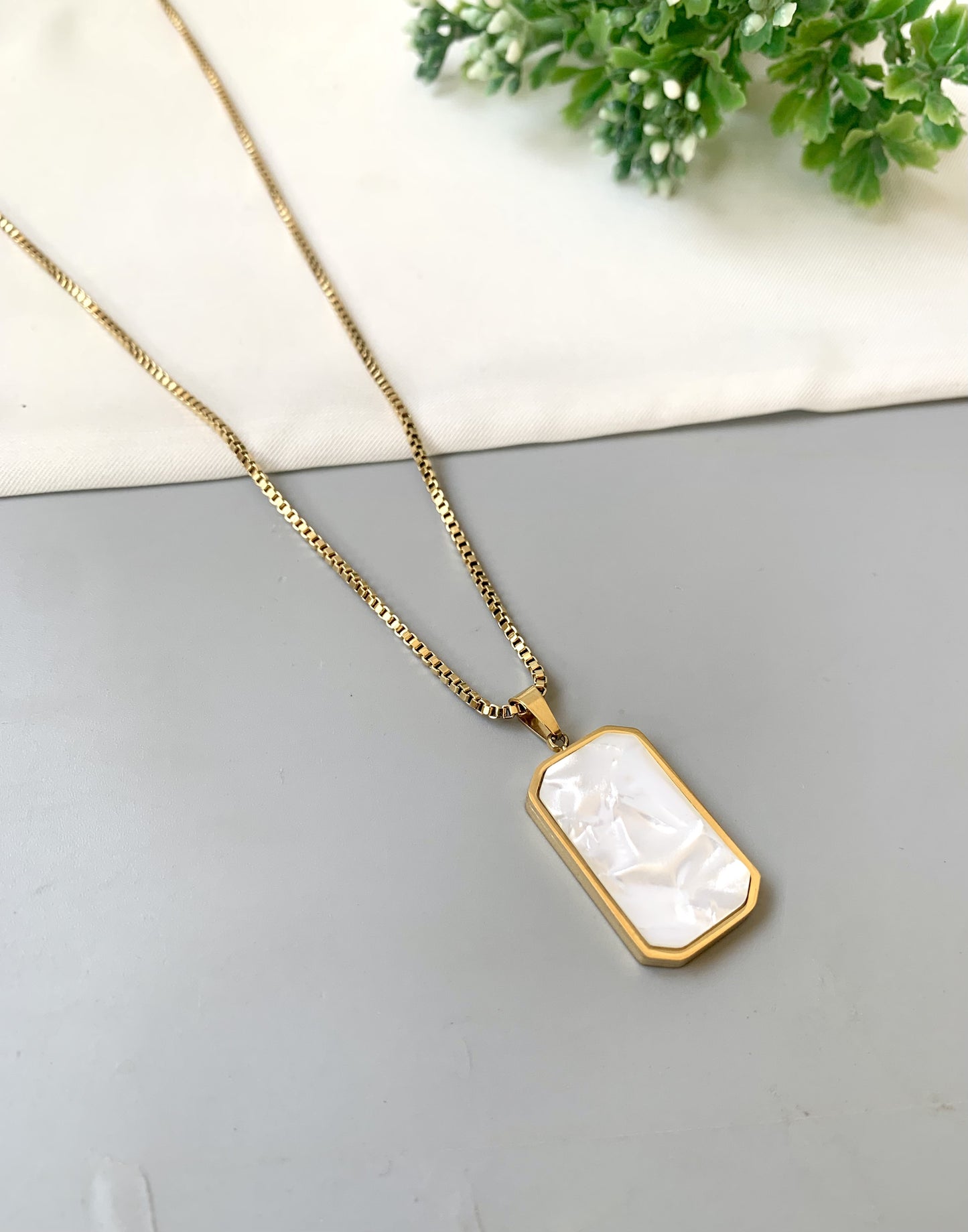 Gold necklace with a rectangular pendant on a gray surface with greenery in the background