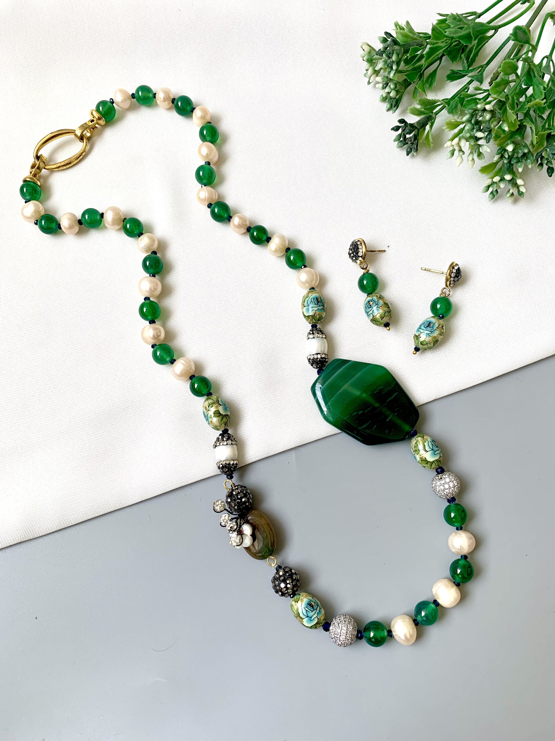 Beaded necklace and earrings with green and white stones on a white surface with greenery.