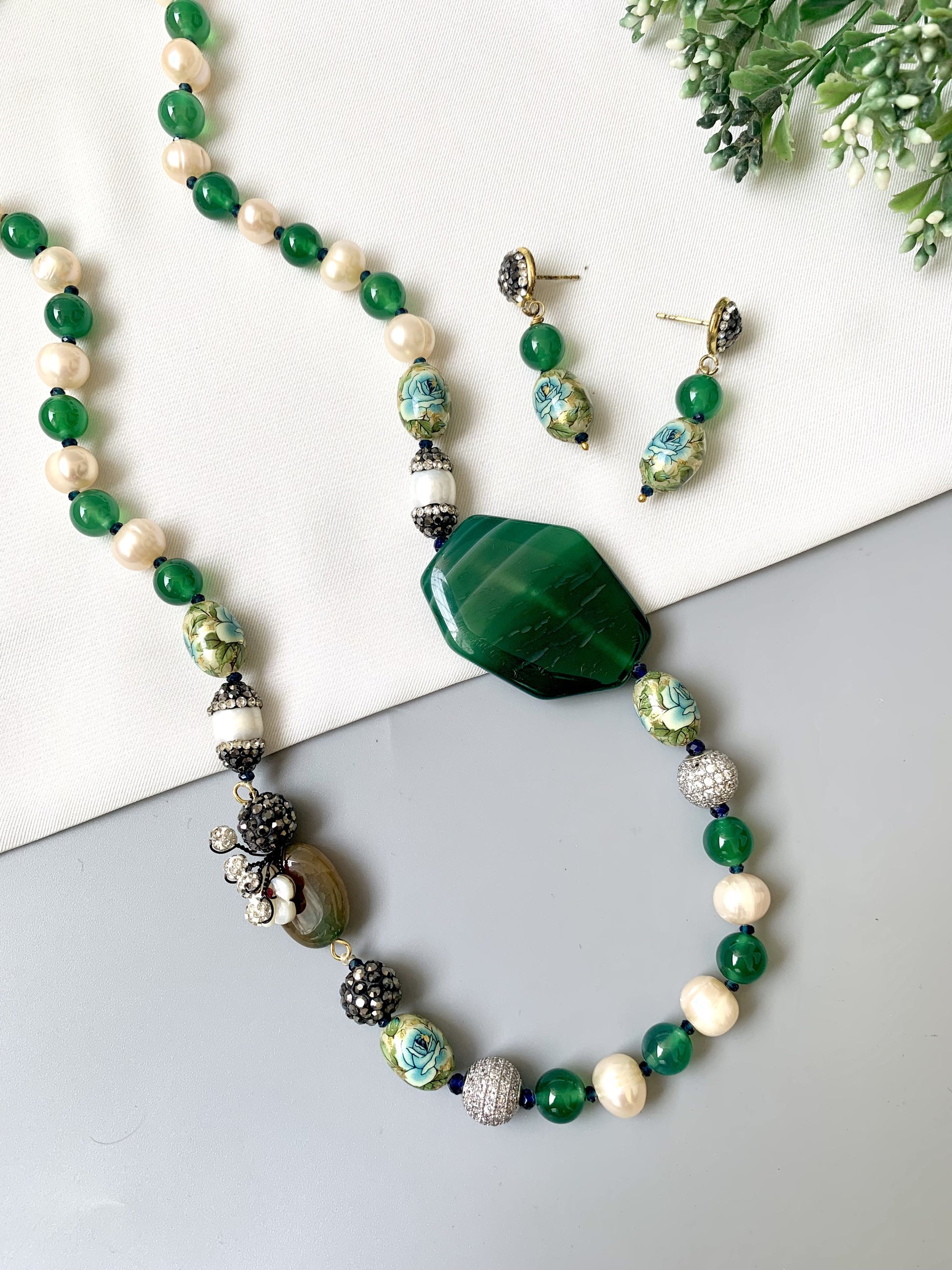 Green and white beaded necklace with a large green stone on a white surface with earrings and flowers.