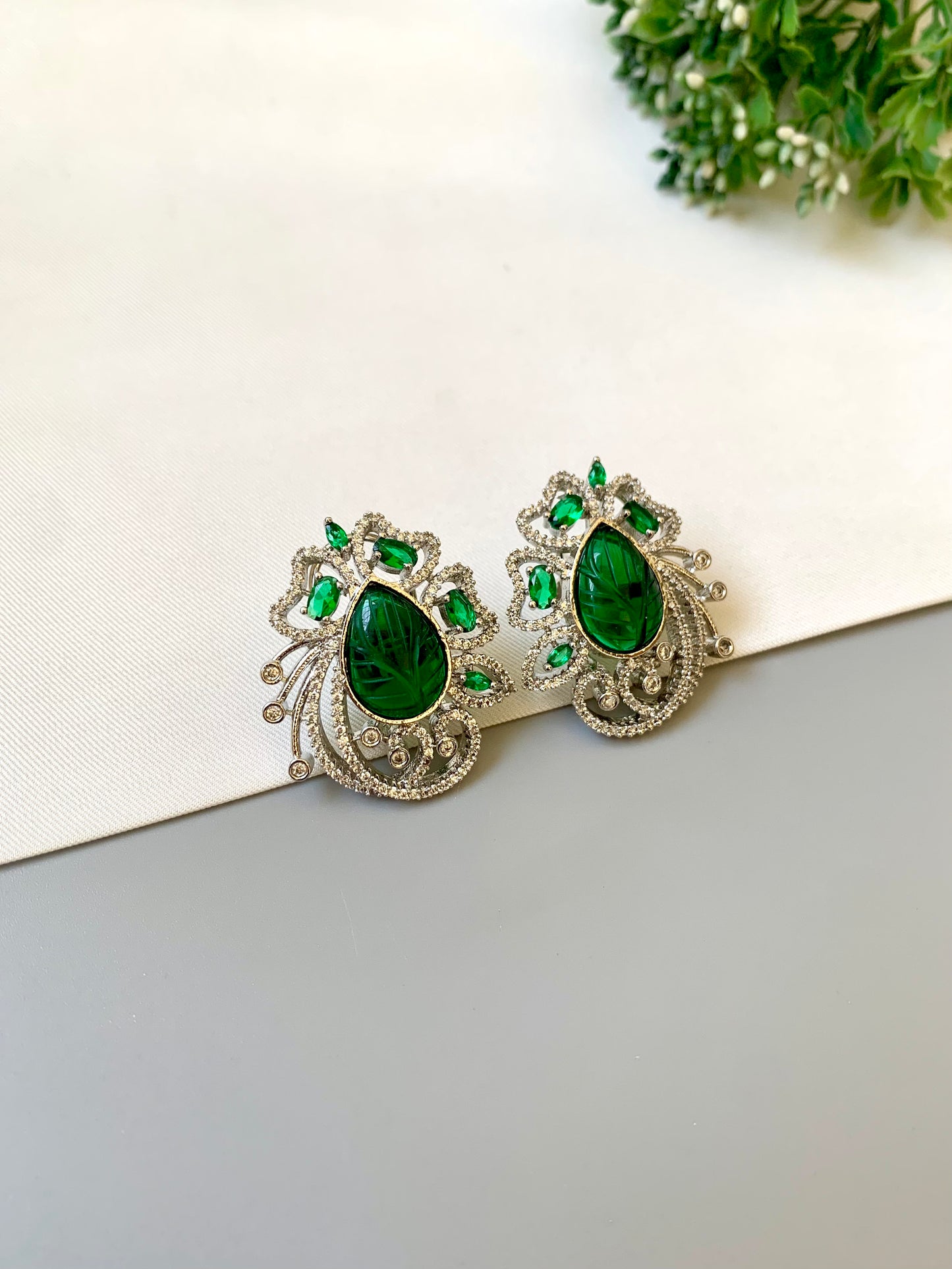Pair of green gemstone earrings on a white surface with a blurred green plant in the background.