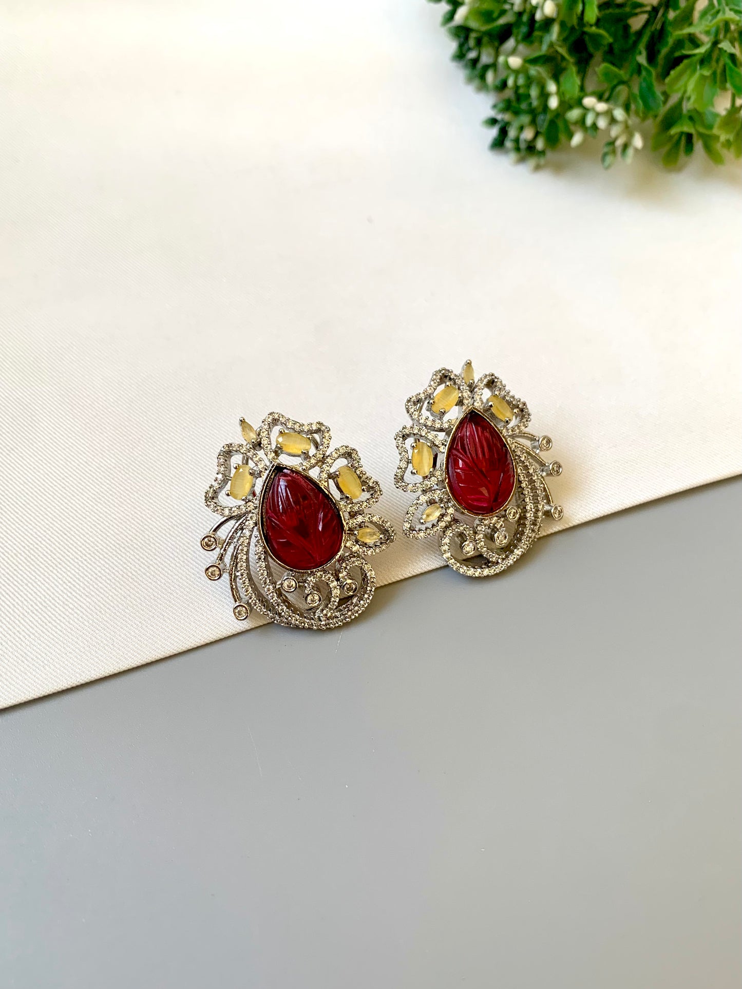 Pair of earrings with red gemstones on a white surface with greenery in the background
