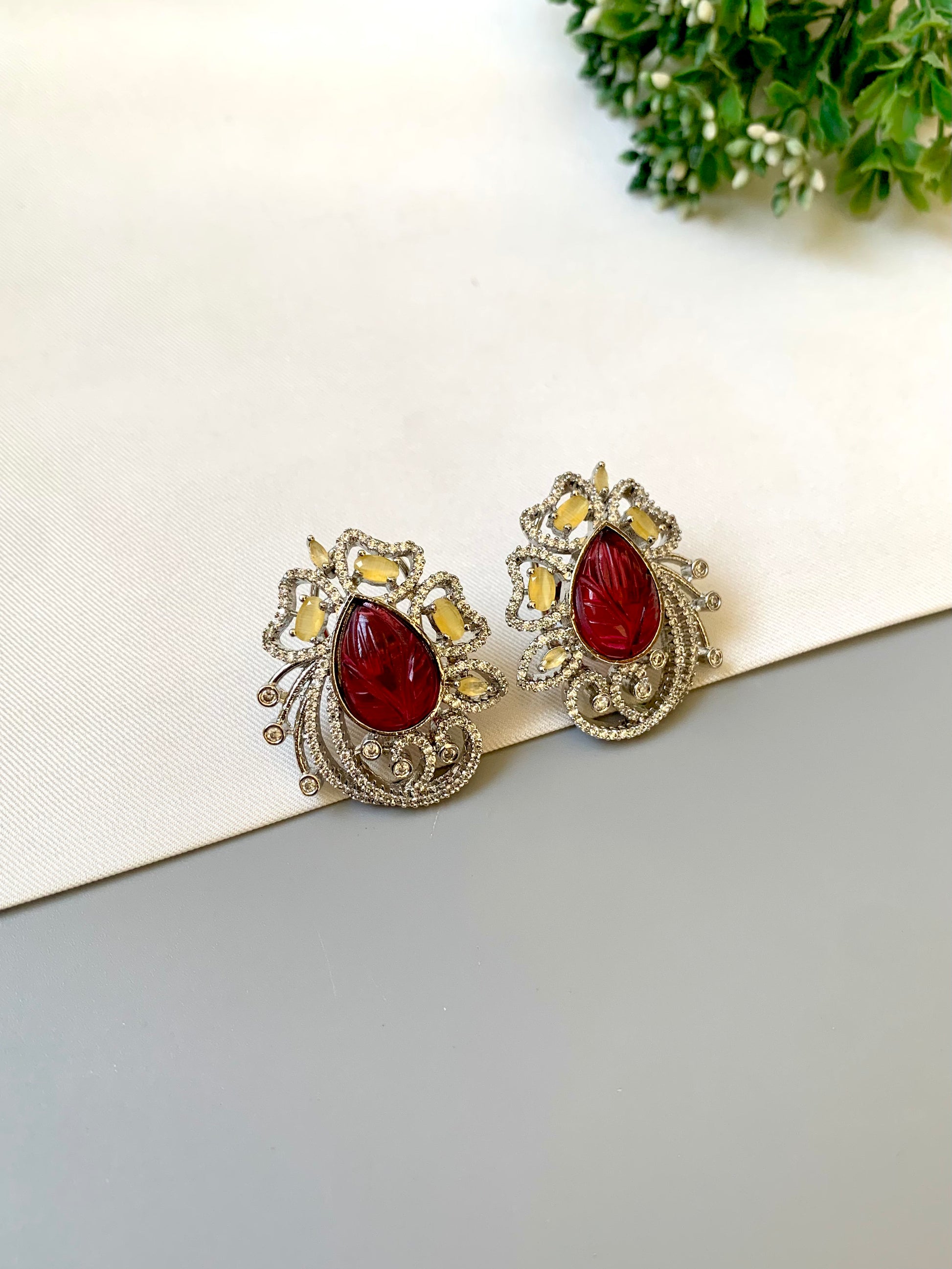Pair of earrings with red gemstones on a white surface with greenery in the background