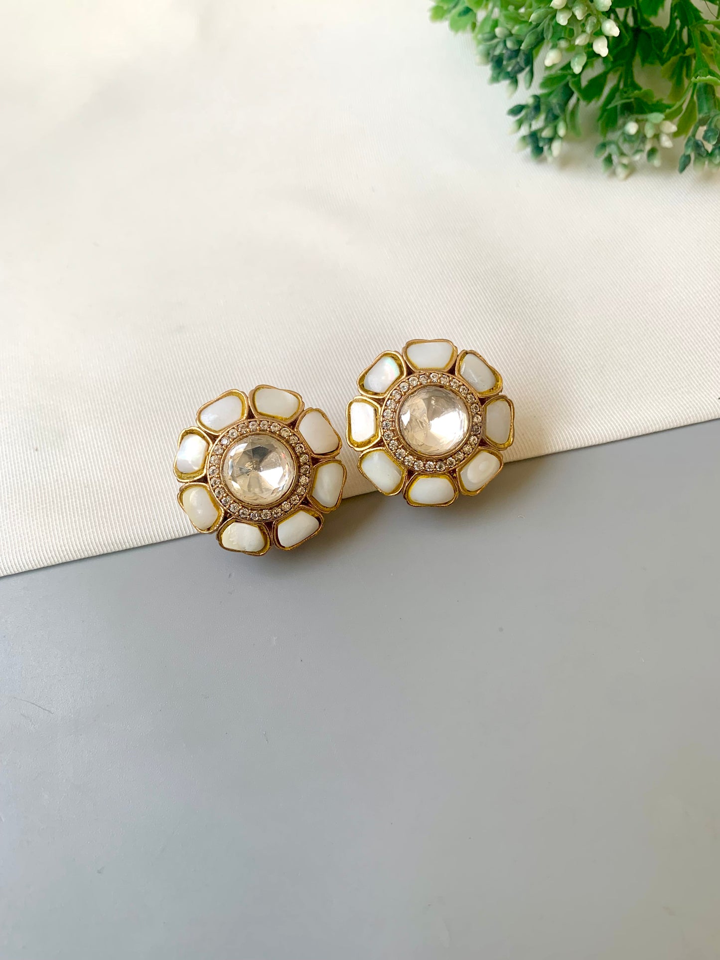 Pair of gold and white earrings on a light gray surface with a white cloth and greenery in the background.