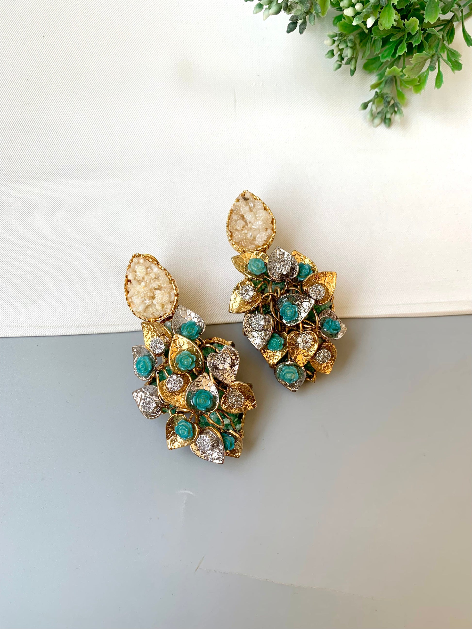 Gold and green gemstone earrings on a light gray surface with a white and green plant in the background.