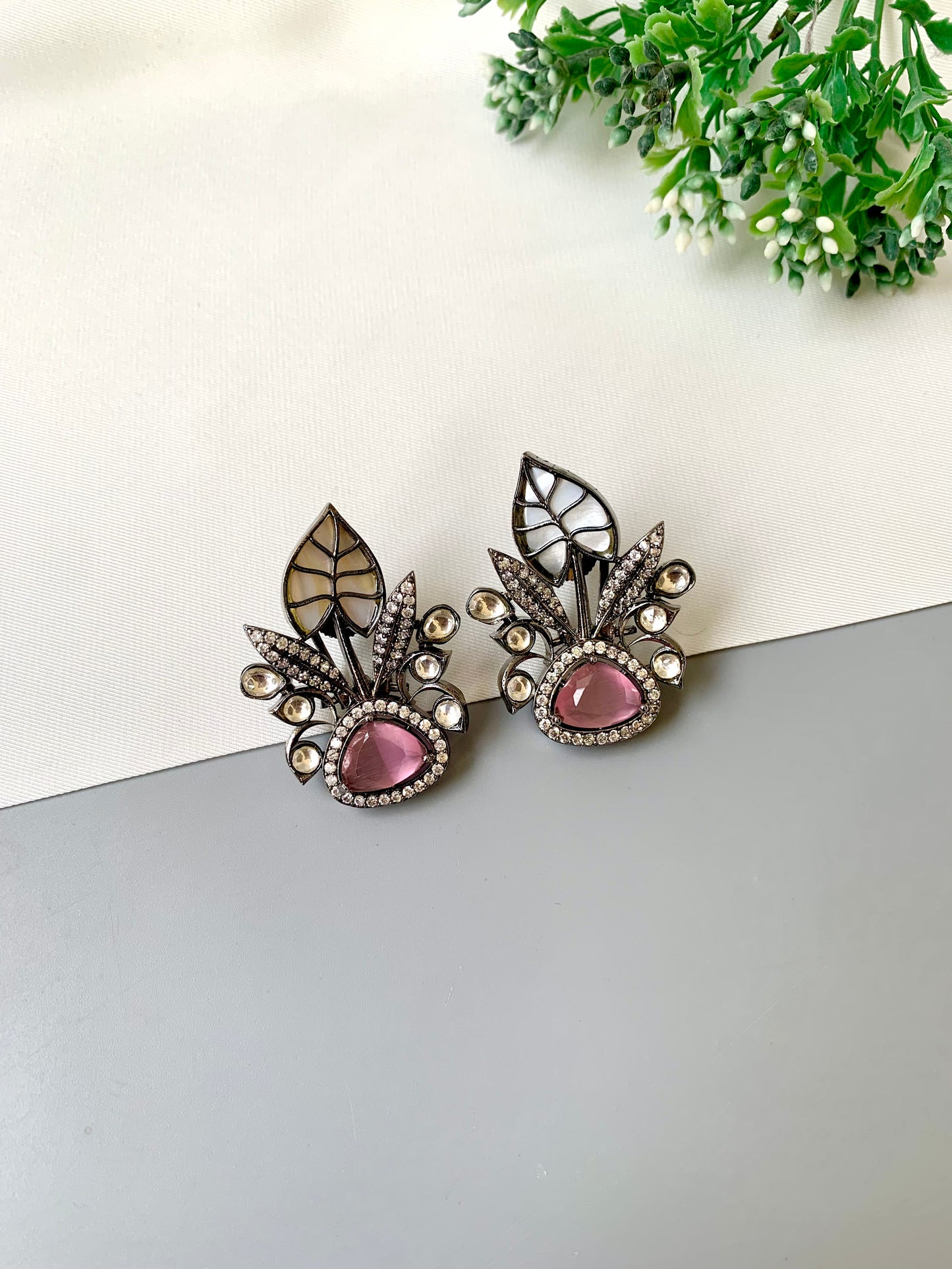 Pair of ornate earrings with pink gemstones on a light surface with greenery in the background