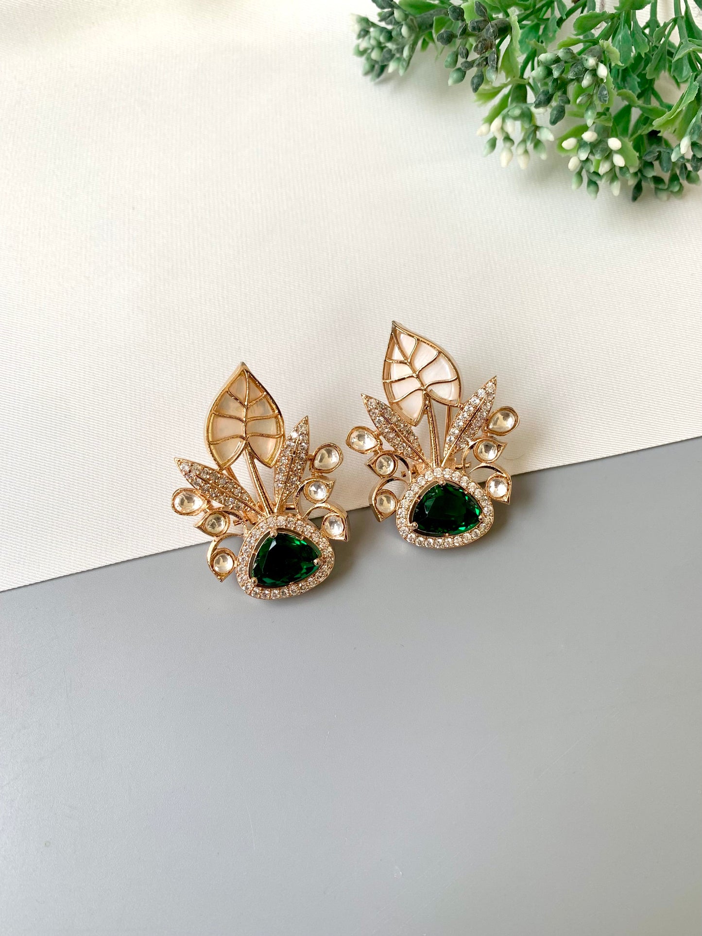 Gold earrings with green gemstones on a white surface with greenery in the background