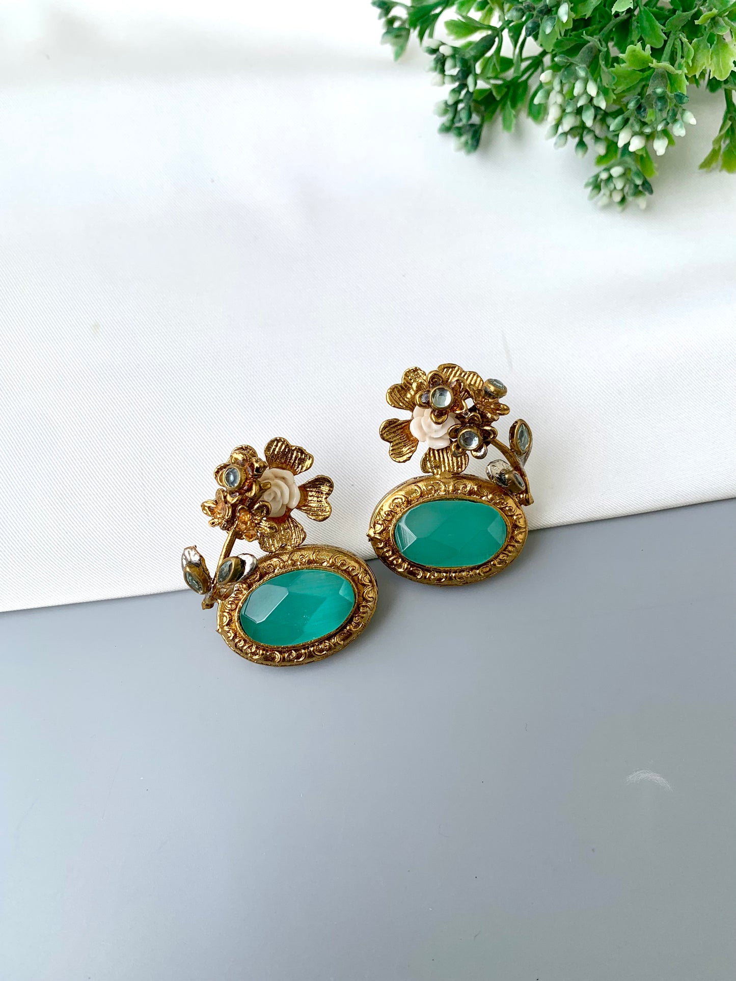 Gold and turquoise earrings on a white surface with greenery in the background